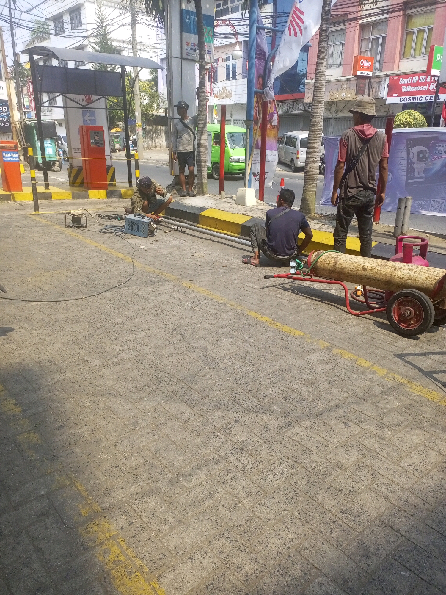 The image depicts an outdoor scene in Bandar Lampung, Indonesia, likely on a street or walkway adjacent to a building. In the foreground, a person is crouched down, appearing to be working on or near a welding machine labeled "LAKONI 200 A". The welding machine is connected to power cables. Nearby, a large wooden log is mounted on a red wheeled cart, with a pink gas cylinder and pressure gauges attached. Another person is seated on the curb near this setup, facing away from the camera. Further back, a person wearing a hat stands near a palm tree, also facing away. In the background, there are buildings, a green vehicle, and various signs, including one for "OLike COSMIC CELL" and another with Indonesian text. The ground is paved with interlocking bricks. The overall scene suggests a work in progress, possibly related to construction or maintenance.