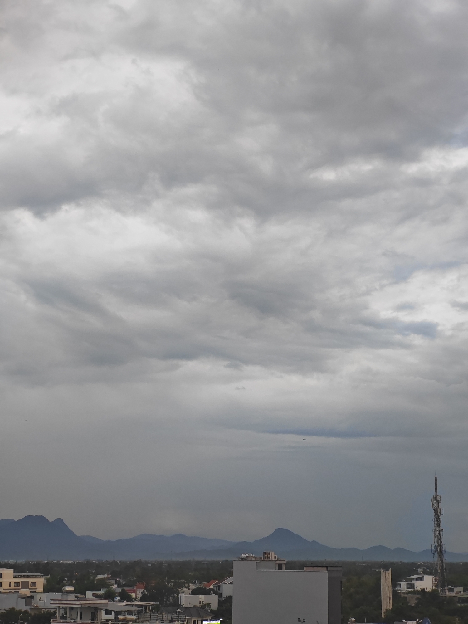 The image captures a panoramic view of a city on a cloudy day, likely in Da Nang, Vietnam, with a range of mountains in the background. The foreground is dominated by the rooftops of several buildings, including a prominent white structure on the right with a tall communication tower. A smaller, light-colored building with visible windows is located to the left of this tower. In the mid-ground, a sprawling urban landscape with a dense collection of buildings and trees is visible. The far background features a chain of layered, blue-hued mountains. High in the sky, a faint airplane can be spotted. The overall atmosphere is overcast, with heavy grey clouds obscuring the sky, suggesting a potential for rain.