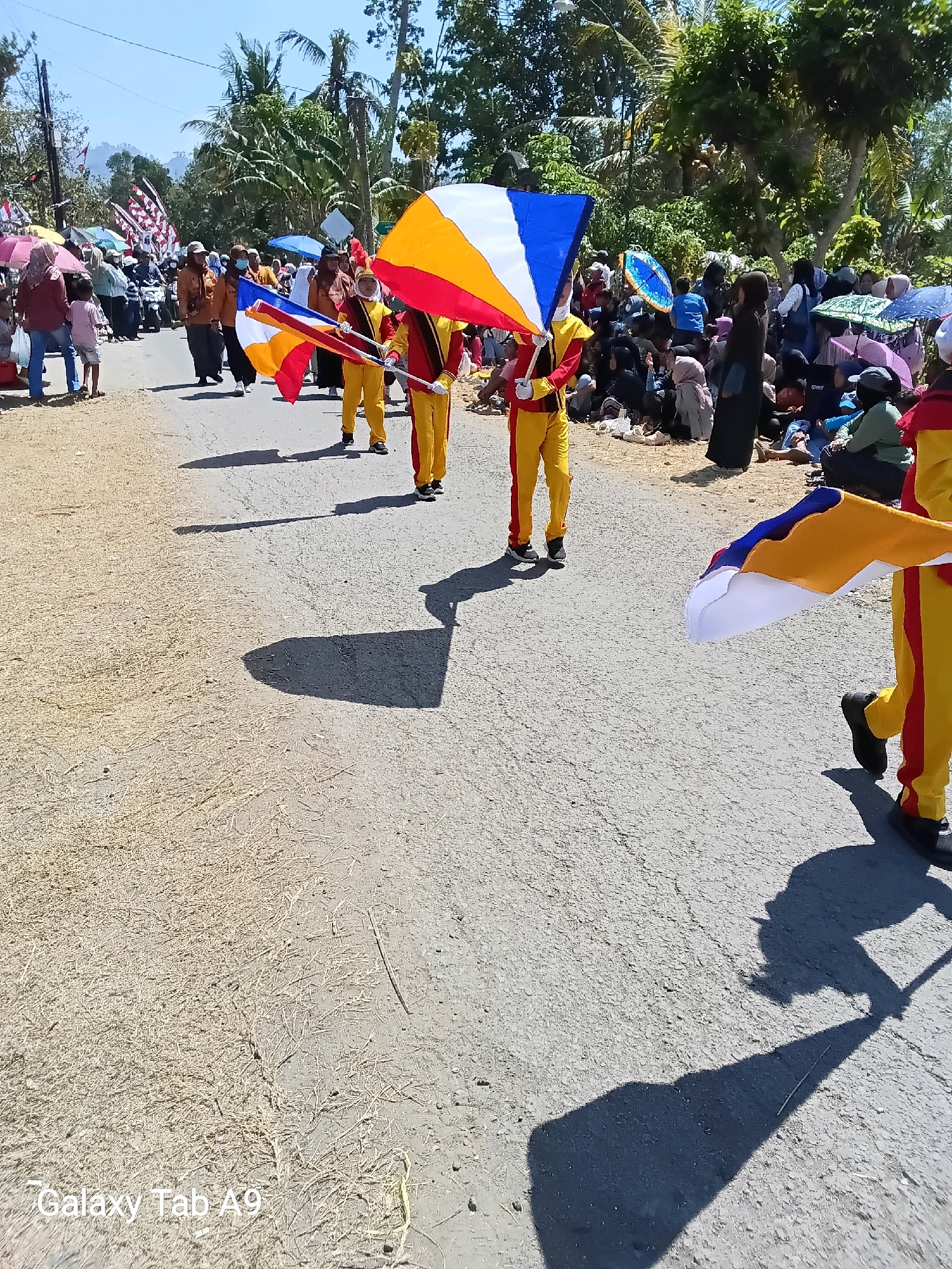 A parade is taking place on a sunny day in the city of Ngadirenggo, Indonesia. Several individuals in yellow and red marching band uniforms are visible, holding colorful flags. One individual in the foreground is holding a flag with white, blue, yellow, and red sections. Another flag, mostly white with yellow and red accents, is visible to the right. The parade route appears to be a paved road with a sandy shoulder covered in dry grass. Spectators are lining the road, some sitting and others standing, with a few umbrellas visible for shade. Trees and foliage are present in the background, suggesting a rural or semi-rural setting within the city. The scene suggests a festive community event.
