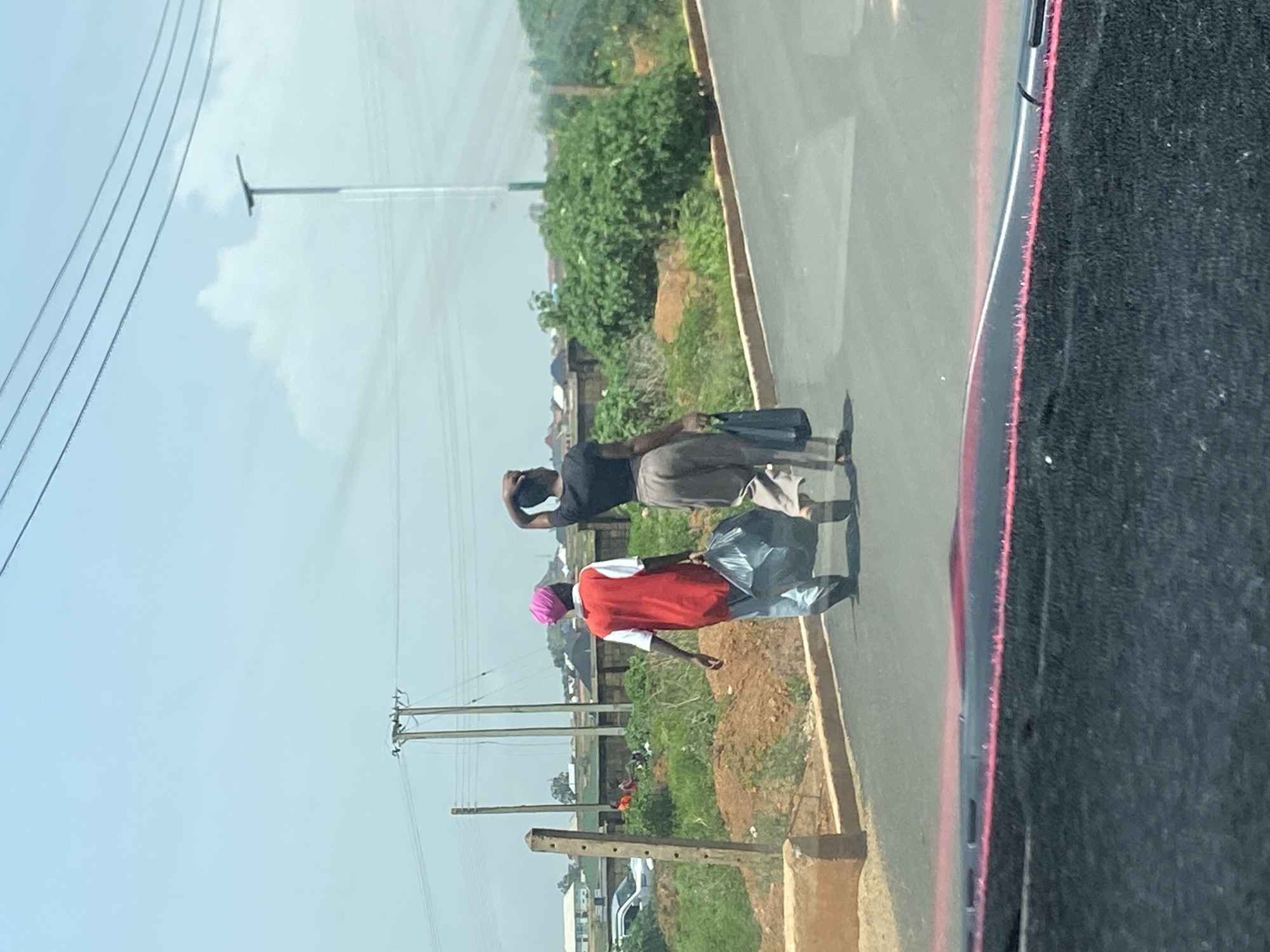 The image shows two individuals standing by the side of a road, with a grassy area and some buildings in the background. The sky is bright and appears to be a clear day, suggesting midday. The road surface is asphalt, and there is a concrete curb or barrier along the edge. Overhead power lines are visible, indicating an urban or semi-urban environment.
One person is wearing a black t-shirt and light-colored pants, carrying a dark bag. The other individual is wearing a red shirt with white accents and a pink head covering, and is holding a large, grey plastic bag. The people appear to be standing still, perhaps waiting for transportation or resting. The scene is consistent with a roadside in Bar Jirgi Summa, Nigeria. There is no visible text in the image.
