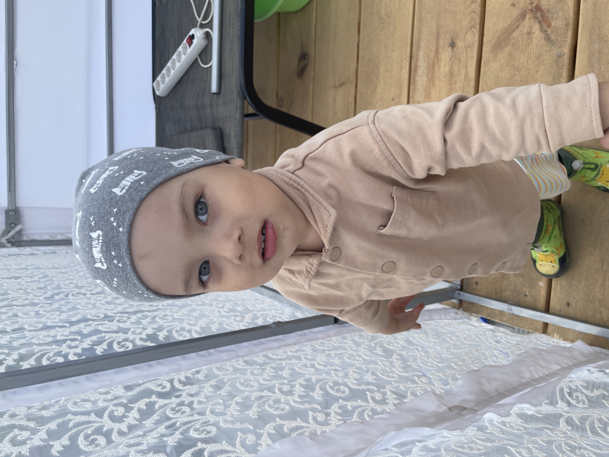 A toddler is standing on a wooden deck covered in white lace fabric. The child is wearing a gray hat, a beige shirt, and striped pants, with green shoes visible. Behind the child is a metal structure covered in the same white lace fabric. A black table with a green object and a white power strip on it can be seen in the background. The scene appears to be outdoors. I have no information about Zav'yalovo, Russia, so I cannot pinpoint the exact location.