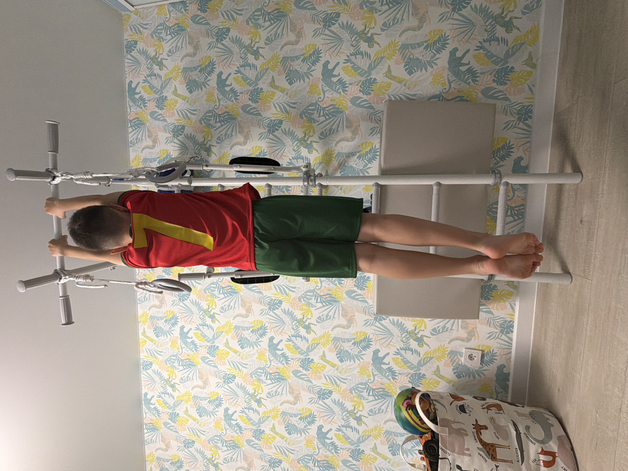 This media file, taken in Votkinsk, Russia, shows a young child hanging from a white Swedish wall or climbing frame. The child, likely a boy, is seen from behind, wearing a red t-shirt with a large yellow number '7' on the back, and dark green shorts. He is gripping an upper horizontal bar of the exercise equipment with both hands, with his body extended downwards.
The wall behind the child is covered with a decorative wallpaper featuring a playful pattern of light blue, yellow, and light pink foliage, alongside stylized animal silhouettes such as monkeys, dinosaurs, and alligators. A white electrical outlet is visible on this patterned wall.
The floor is made of light-colored wooden planks, bordered by a white baseboard. In the lower right corner of the image, a fabric storage basket is partially visible. This basket is adorned with illustrations of various cartoon animals, each labeled with its English name, including "ELEPHANT," "HIPPO," "GIRAFFE," "ALLIGATOR," and "DINOSAUR." A colorful ball can be seen inside the basket. The scene depicts an indoor setting, likely a child's room or play area, illuminated by indirect light. The time of day is not discernible.