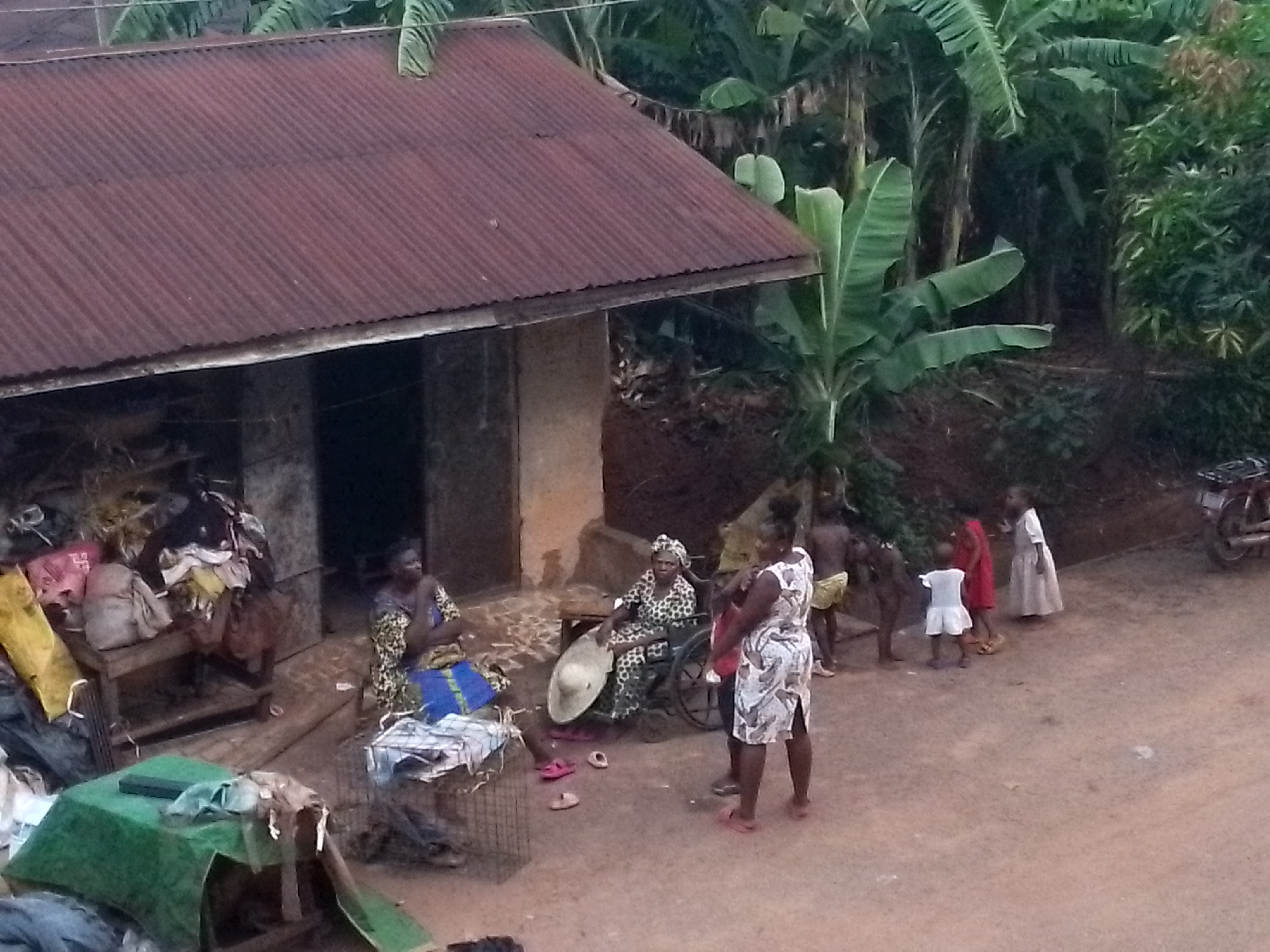 The image shows a scene in Orlu, Nigeria, featuring a corrugated metal roof house with two open doorways. In front of the house, three adult women are present. One woman sits on a chair with a blue cloth, another sits in a wheelchair holding a straw hat, and a third stands talking to them wearing a patterned dress. Nearby, a group of children in various states of dress are playing. A motorcycle is parked in the background. The environment includes banana trees and other vegetation, suggesting a rural setting. There is clothing and other items piled on a bench near the house. The ground is dirt.