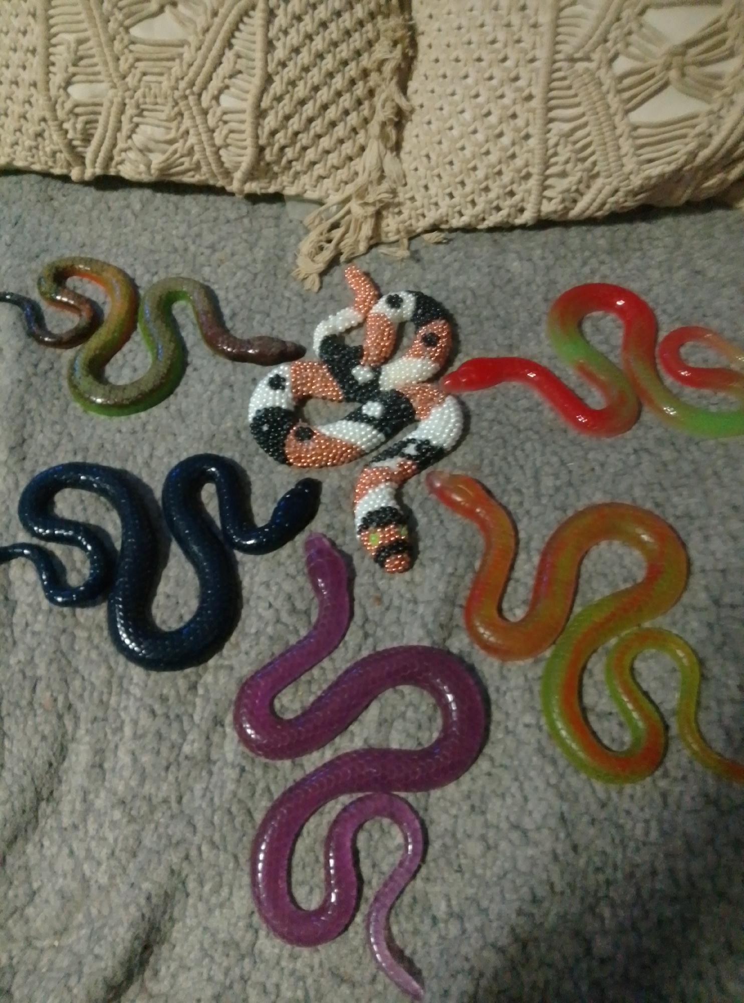 The image displays a collection of toy snakes arranged on a grey, textured surface. A cream-colored, macrame-style fabric object is visible in the upper portion of the frame. The snakes are of various colors and materials, including solid colored, translucent, and beaded snakes. They are positioned in a scattered, winding formation across the surface. There are no people or animals present in the image. The scene is indoors, with no discernible view of the city of Wedel, Germany.