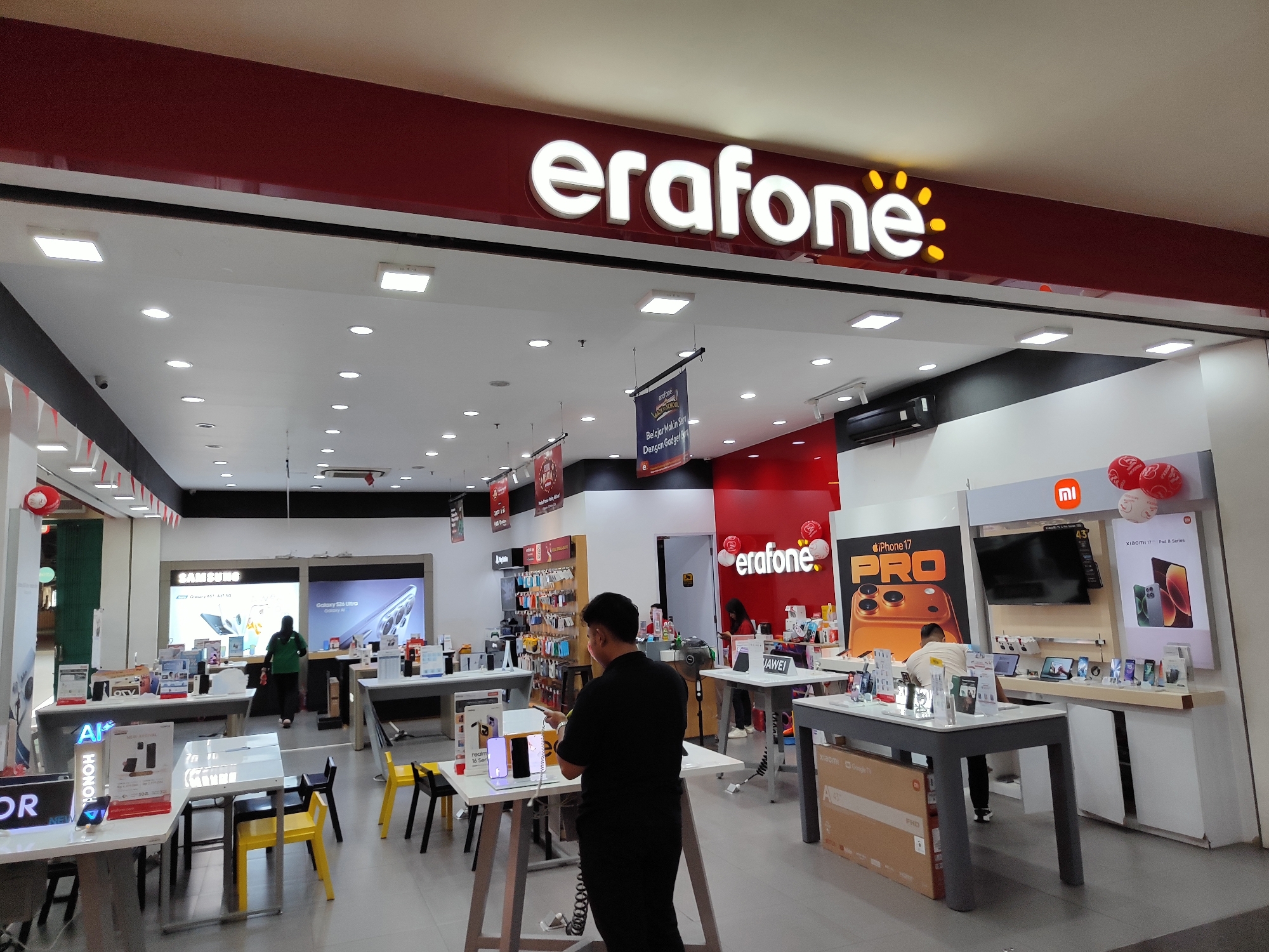 This is a medium shot of the interior of an Erafone store, likely located in Sempusari, Indonesia. The store is brightly lit with ceiling lights and displays featuring various electronic devices. A large Erafone logo with illuminated white text and yellow accents is prominently displayed above the entrance.
In the foreground, there are several display tables showcasing mobile phones and other accessories. One table in the center has a person in a black shirt interacting with a display of smartphones. Yellow and black chairs are arranged around some of these tables. To the left, a display table has "HONOR" branding.
Further back, a Samsung display features images of their latest phones. A woman in a green uniform is visible behind a counter, attending to a display. On the right side of the store, there are displays for iPhones and Xiaomi devices, including a large screen showing a promotional image for "iPhone 17 PRO". Another person, wearing a light-colored shirt and dark pants, is examining items at a display table on the right.
The store environment appears clean and organized, typical of a retail electronics store. Red and white banners or flags are visible hanging from the ceiling in the background. Balloons, some with Erafone branding, are also present on the right side. The overall impression is of a modern and well-stocked mobile phone retail outlet.
