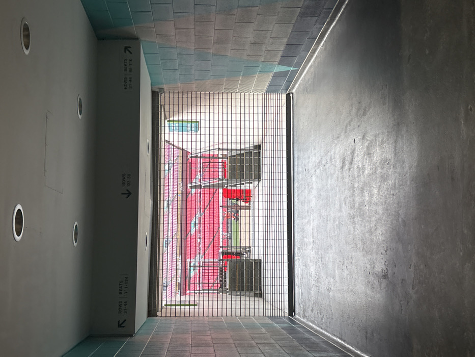 This media file captures an interior view from a stadium concourse or hallway, looking out onto the seating area and pitch. The perspective is from a relatively dim corridor that opens into the brighter stadium bowl through a black metal grated barrier.
On the left side of the corridor, a tall, smooth grey wall is visible, featuring several circular recessed lights. This wall displays multiple signs indicating seating directions. The upper, more distant sign reads "ROWS SEATS 31-44 95-110" with an arrow pointing right, towards the stadium. Closer to the viewer, another sign reads "ROWS 02-30" with an arrow pointing left. A third sign, partially visible further left, indicates "ROWS SEATS 31-44 111-124" with an arrow pointing left. The corridor floor is composed of dark grey and greenish-blue rectangular tiles. The ceiling in the upper background (left side) features light blue and grey rectangular tiles. The right side of the corridor is a textured, dark grey concrete wall.
Through the metal grating, the vibrant interior of a stadium is revealed. Numerous rows of bright red seats are arranged in tiers, descending towards a green sports pitch, a section of which is visible at the lowest level. Support structures for the seating and a small, greenish-tinted window are also visible in the distance. The stadium is empty, with no people present. The lighting indicates it is daytime. Based on the provided context, this scene is set within Wembley Stadium in the United Kingdom.