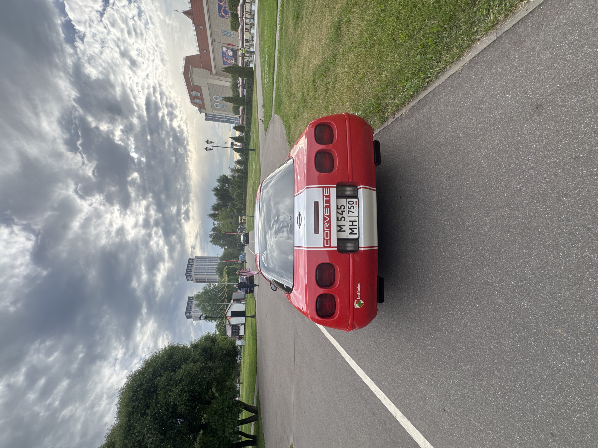 This image captures the rear view of a red and white Chevrolet Corvette parked on an asphalt path next to a grassy area. The car has a visible license plate that reads "M 545 MH 750 RUS". The backdrop features a large, ornate building with red roofing, a park with trees and green grass, and in the distance, tall modern buildings. The sky is filled with dramatic, cloudy formations, suggesting an overcast or partly cloudy day.
The overall scene appears to be an outdoor event or gathering, possibly a car show or a public park with some form of exhibition. There are several indistinct figures of people visible in the background, some near the main building and others near smaller structures within the park. The lighting indicates it could be daytime, likely afternoon, given the brightness of the sky and reflections on the car. The presence of the Russian license plate and the architectural style of the distant buildings hint at the location being Moscow, Russia. The image has a slightly tilted perspective, giving a dynamic feel to the composition.