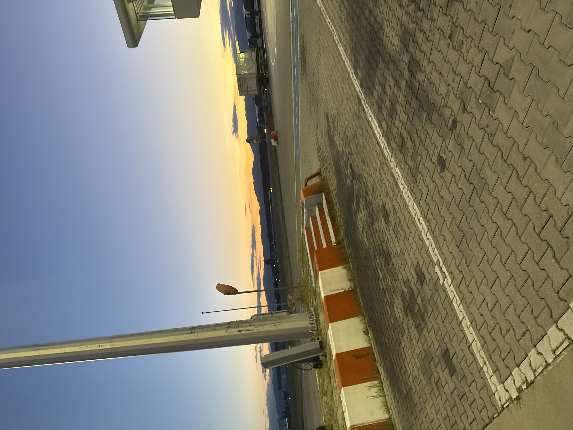 This is a photograph of an airport tarmac at sunset, likely at Sofia Airport, Bulgaria. A large, metallic pole stands prominently on the left side of the frame, casting a shadow onto the interlocking concrete pavers that make up the ground. In the middle ground, a series of white and orange concrete barriers are visible, some with a stepped design. Beyond the barriers, several buses and a large refrigerated truck are parked. In the distance, mountains form a silhouette against a sky transitioning from a deep blue to a warm orange and yellow gradient, indicative of sunset. A few aircraft can be seen on the runway in the far distance. A small building with windows, possibly a control tower or airport facility, is visible on the upper right. The overall scene suggests a quiet, operational moment at the airport as day turns into night.