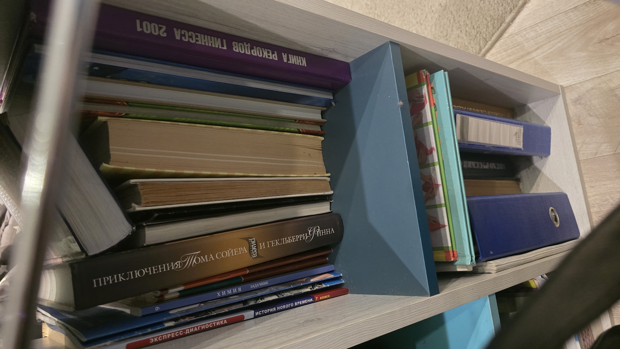 The image shows a bookshelf filled with various books and binders. On the top shelf, there are several books with their spines facing outwards. One book has Russian text that appears to read "Приключения Тома Сойера" (The Adventures of Tom Sawyer). Another book has the title "КНИГА РЕКОРДОВ ГИННЕССА 2001" (Guinness World Records 2001). Below this shelf, there is another shelf with what appears to be binders and more books. One blue binder is prominently visible. The bookshelf itself is made of light-colored wood with a blue shelf insert. The surrounding environment is not clearly visible but appears to be an indoor setting with a carpeted floor. There are no people or animals depicted in the image. The scene is likely a domestic or office setting, the exact location in Ukhta, Russia, is unknown.