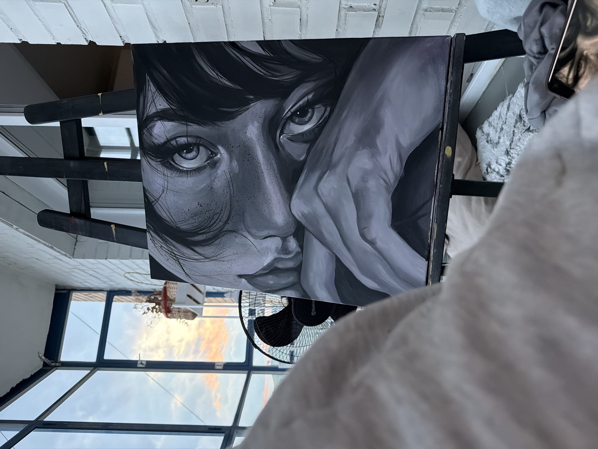 The image depicts an indoor scene with a painting on an easel in the foreground. The painting is a monochrome artwork of a person's face, focusing on their eyes and hands. The background includes a window with a view of a cloudy sky with hints of sunset colors, a black fan, and a white brick wall. The overall setting appears to be a balcony or a room with a view. There are no identifiable landmarks or contextual clues within the image to suggest a specific location within Krasnoyarsk, Russia.