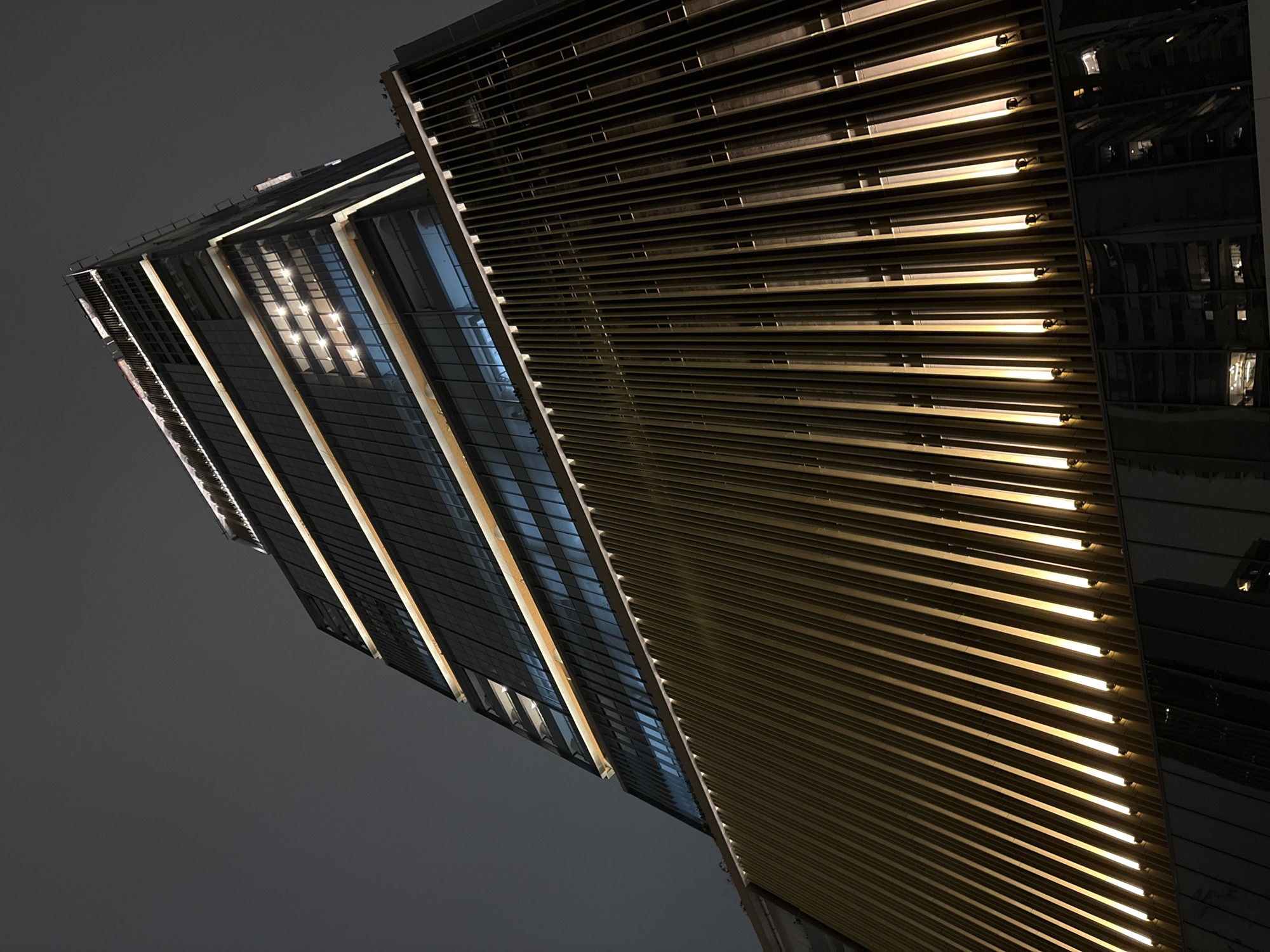 The image shows a low-angle, nighttime exterior view of a modern skyscraper in São Paulo, Brazil. The building features a distinctive design with alternating bands of dark glass and illuminated horizontal strips of warm light. The lower portion of the building is clad in a vertical slatted gold-colored facade, with upward-facing lights creating a dramatic effect. The sky is dark and overcast, indicating night or poor weather conditions. The reflections in the glass windows suggest that other buildings are present in the vicinity. There are no people visible in the image. The overall impression is one of contemporary urban architecture, illuminated against the night sky.