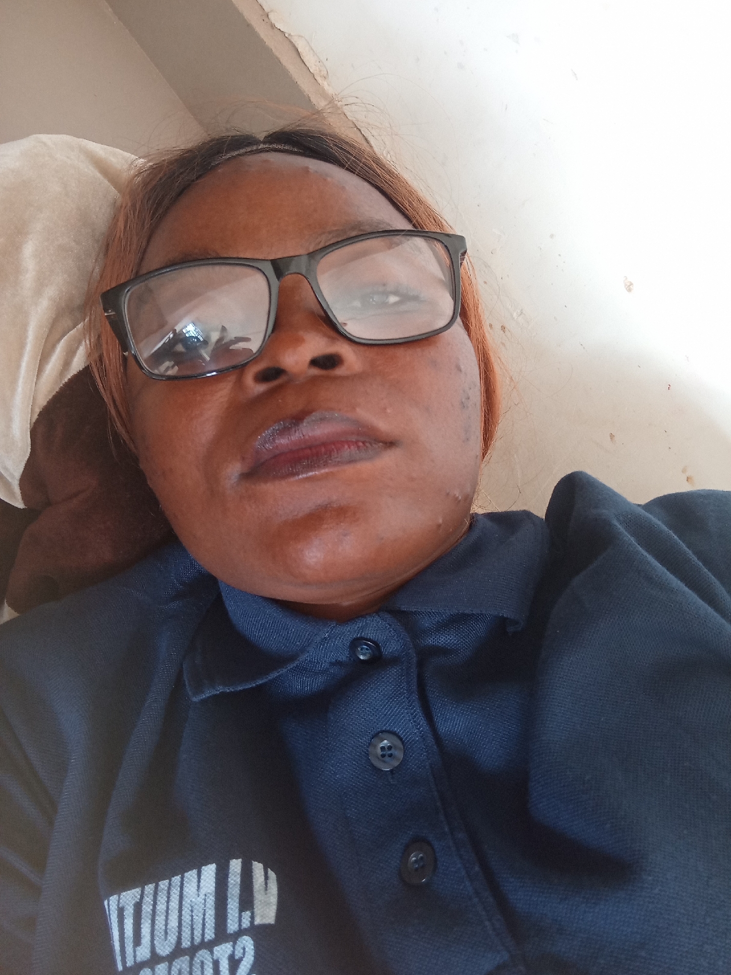 A medium shot captures a woman wearing black-rimmed glasses and a navy blue collared shirt. She is lying down, with her head resting on a beige pillow. Her hair is reddish-brown and pulled back. The background is a plain, light-colored wall with some visible imperfections. Text is visible on the left side of her shirt, but it is partially obscured and difficult to read. The overall lighting is bright, suggesting daytime. The scene appears to be indoors, possibly in a bedroom or a similar personal space.
