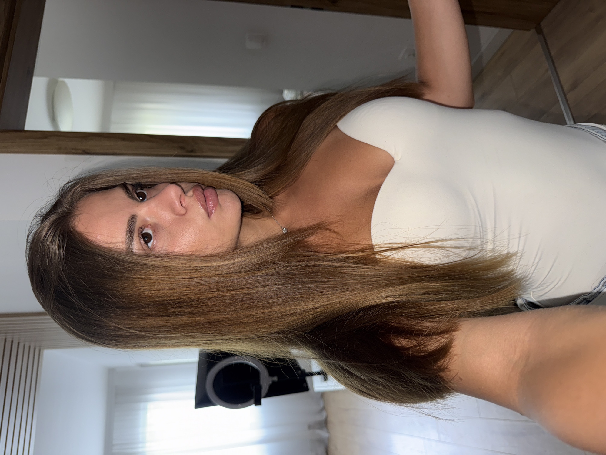 A young Caucasian woman with long, straight brown hair is shown in a medium shot, looking to her right with her mouth slightly parted. She is wearing a white strapless top and a delicate silver necklace. The background appears to be an interior setting, possibly a home or studio. To the left of the woman, a ring light on a stand is visible. The room features white curtains and wooden flooring. The overall lighting is bright, suggesting a well-lit space. There is no indication of the city of Plovdiv, Bulgaria.