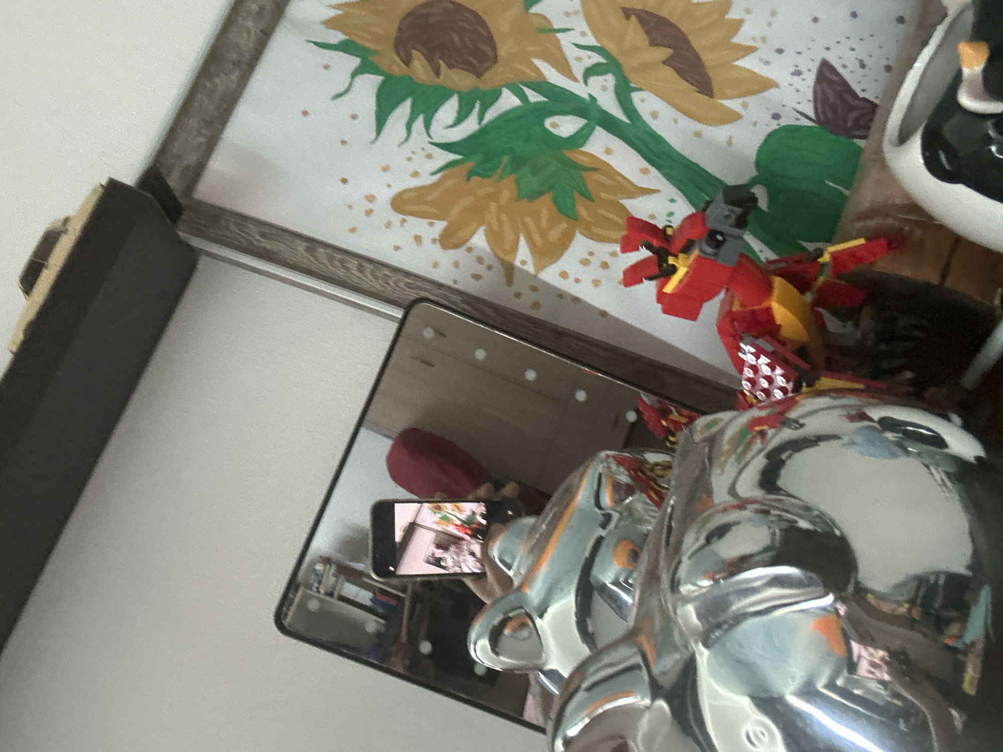 In a room with white walls, a mirror reflects a person holding a smartphone. In front of the mirror, a shiny silver piggy bank is visible. To the right, a painting of sunflowers is displayed. A red and yellow Lego figurine is placed in front of the painting. The scene does not provide enough information to determine a specific location in Plovdiv, Bulgaria.