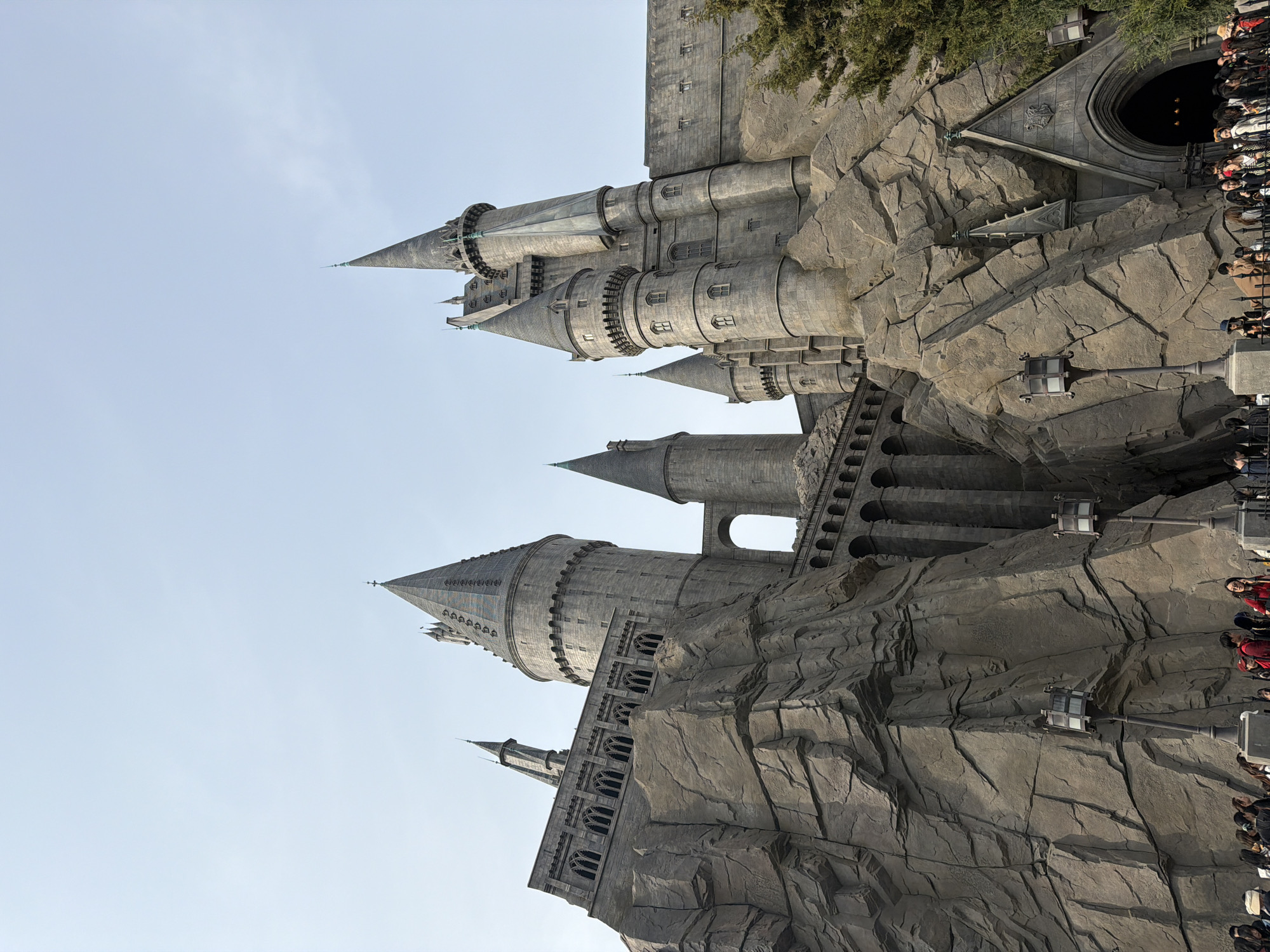 The image features a low-angle, close-up view of Hogwarts Castle, part of the Wizarding World of Harry Potter attraction at a Universal Studios theme park. The castle is constructed of gray stone and exhibits intricate architectural details, including multiple towers, turrets, arched walkways, and crenellations. The castle is situated on a rocky, craggy landscape. A crowd of people is visible on the right side of the frame, suggesting a queue or pedestrian pathway. The sky is clear and pale blue. The scene appears to be an outdoor setting at a popular tourist destination. The location is not in Tongzhou District, China.