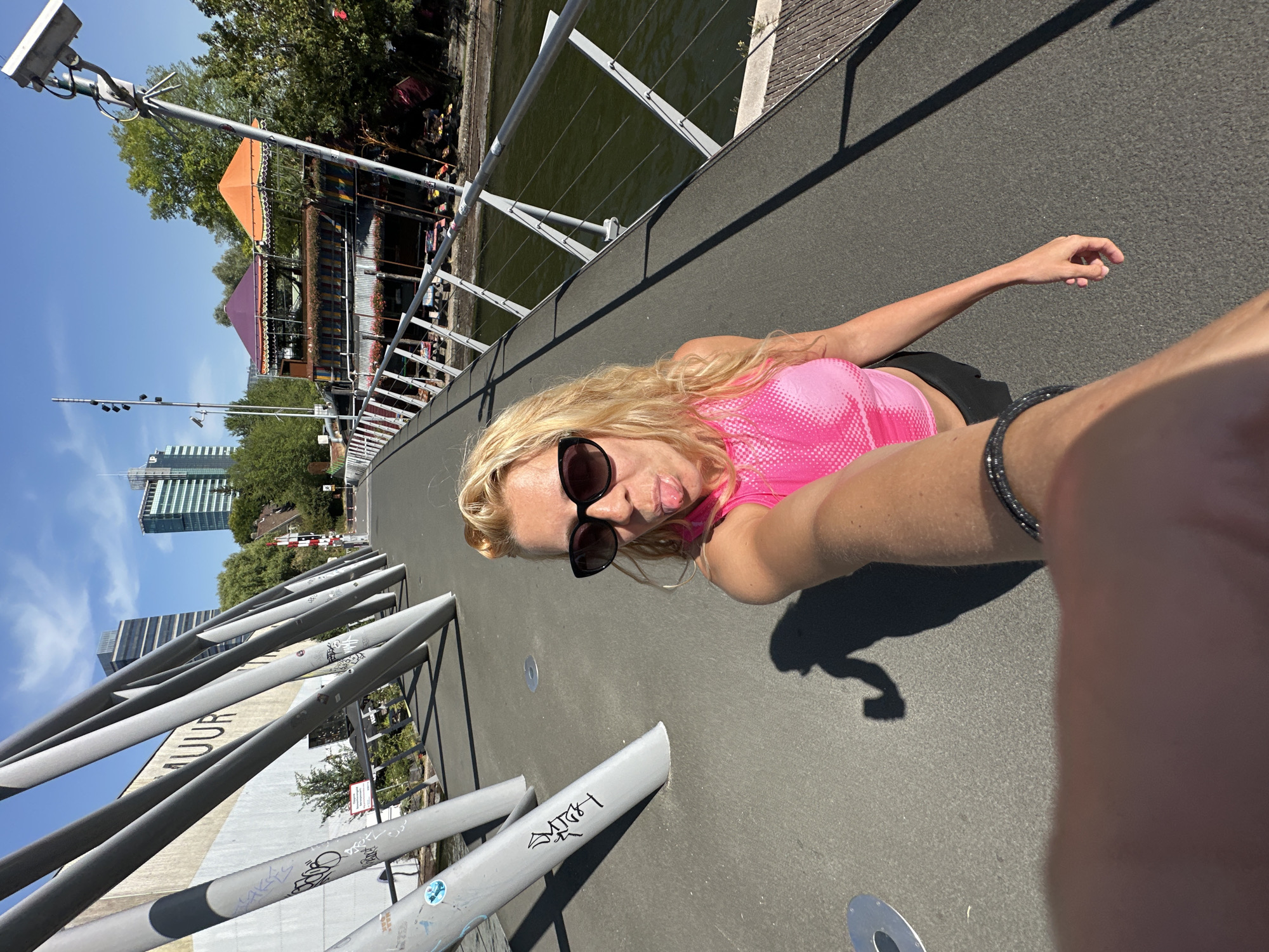 A woman with blonde hair, wearing sunglasses and a pink top, sticks her tongue out for a selfie on a bridge in Amsterdam, Netherlands. The bridge has a modern design with angled metal supports. In the background, there are buildings, trees, and a canal with a boat. The woman's arm, holding the phone, is visible in the foreground.