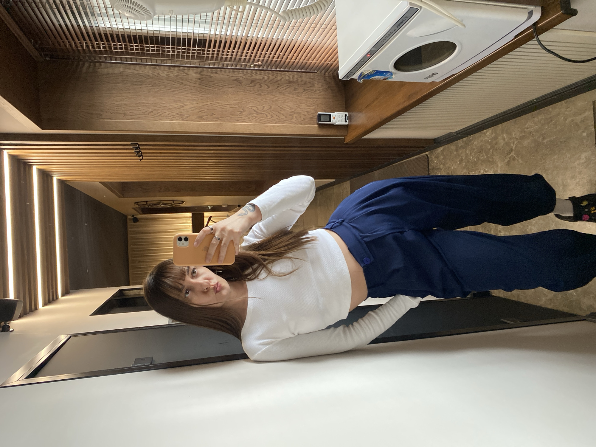 A woman is taking a selfie indoors. She's wearing a white long-sleeved crop top and dark blue pants. She's holding a light orange phone up to take the photo. In the background, there's wood paneling on the walls and ceiling. A washing machine is visible in the upper right corner. Further back, there's a lit hallway. The location or city cannot be determined from the image.
