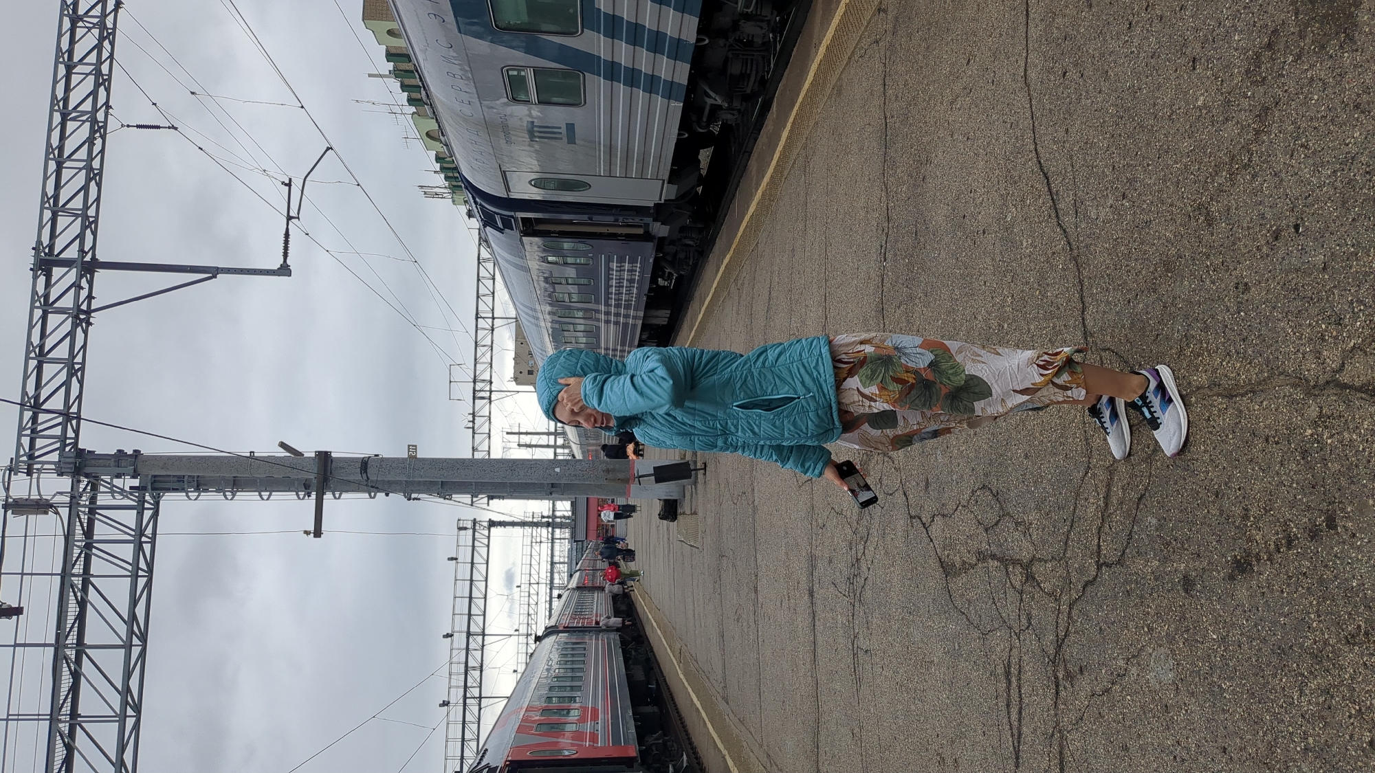 The photograph depicts a person standing on a platform at a train station. The person, who appears to be a woman, is wearing a blue jacket, printed trousers, and athletic shoes. She has her hand up to her head near her hood, and she is holding a smartphone in her other hand. Parallel to the platform on either side are train cars, one with blue and white coloring and the other with red and gray coloring. Overhead support structures, wires, and poles are visible, and the sky is overcast. The location is consistent with a train station in the city of Saratov, Russia.