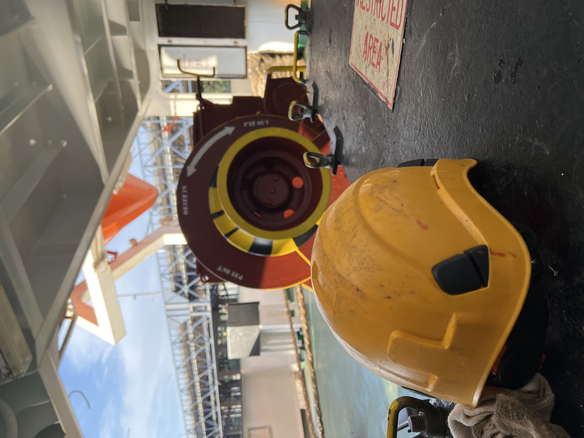 This image captures a yellow hard hat resting on a dark, textured surface, possibly a deck, with a red and yellow winch mechanism in the background. The winch has markings, including arrows and text that appears to say "HEAVE IN" and "PAY OUT." Further in the background, a life raft is visible, along with a metal structure resembling a bridge or walkway. The sky is blue with some clouds, suggesting an outdoor setting. A red and white sign reading "RESTRICTED AREA" is partially visible in the lower right corner. The scene appears to be on a ship or a similar maritime industrial environment. The location is identified as Matosinhos, Portugal.