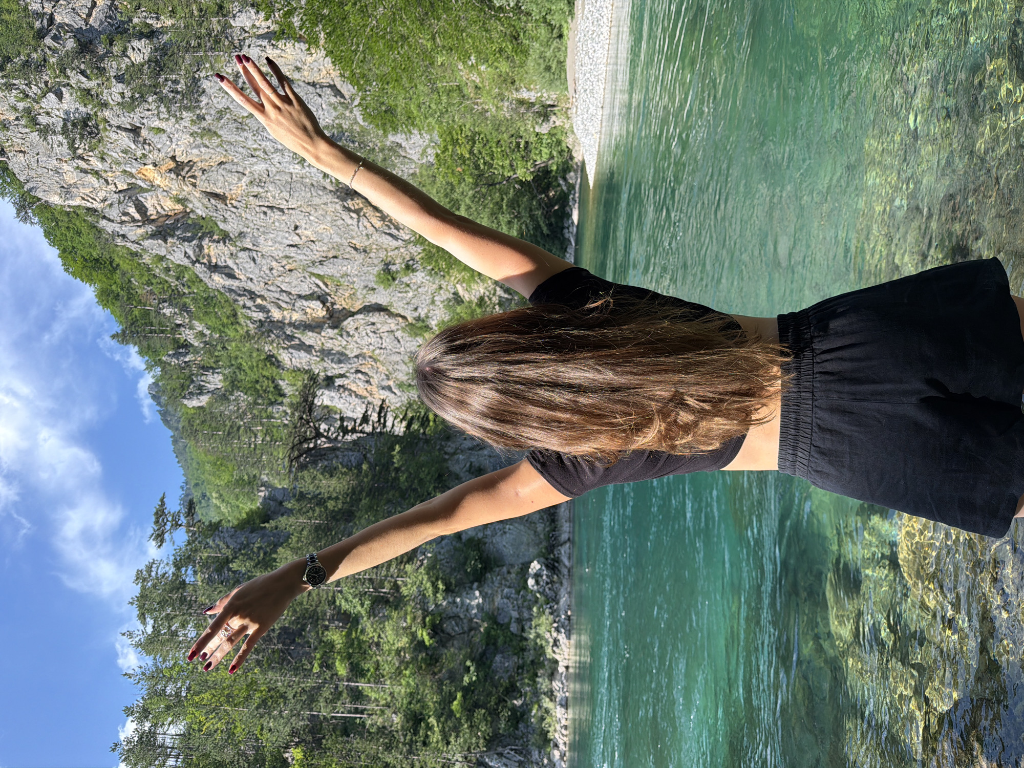 In Bistrica, Montenegro, a young woman stands with her arms raised in celebration near a river. The woman, viewed from the back, has long brown hair and is wearing a black crop top and black shorts. She is adorned with a watch on her left wrist and rings on her fingers. Her right arm displays a bracelet. The river in front of her is a vibrant turquoise, with clear water revealing a rocky bottom. Behind her, a majestic mountain range covered in lush green trees rises, under a bright blue sky with scattered clouds. The scene captures a moment of joy and appreciation for the natural beauty of the area.