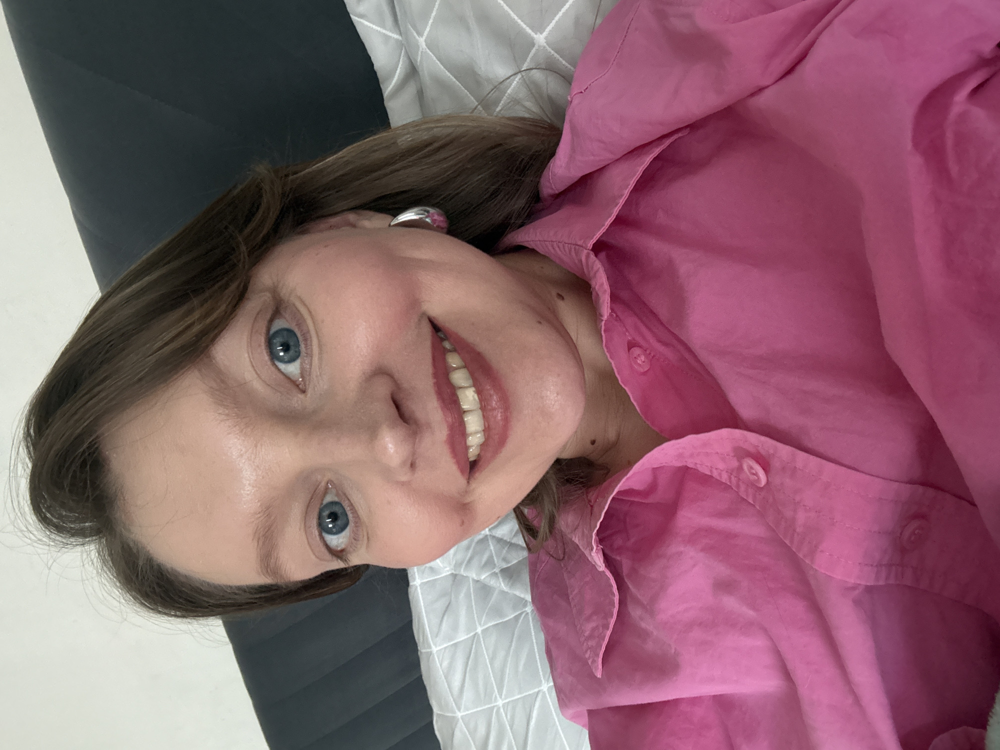A woman lies on a bed, smiling at the camera. She is wearing a pink collared shirt. The bedding includes a gray and white patterned comforter. The location is not discernible.