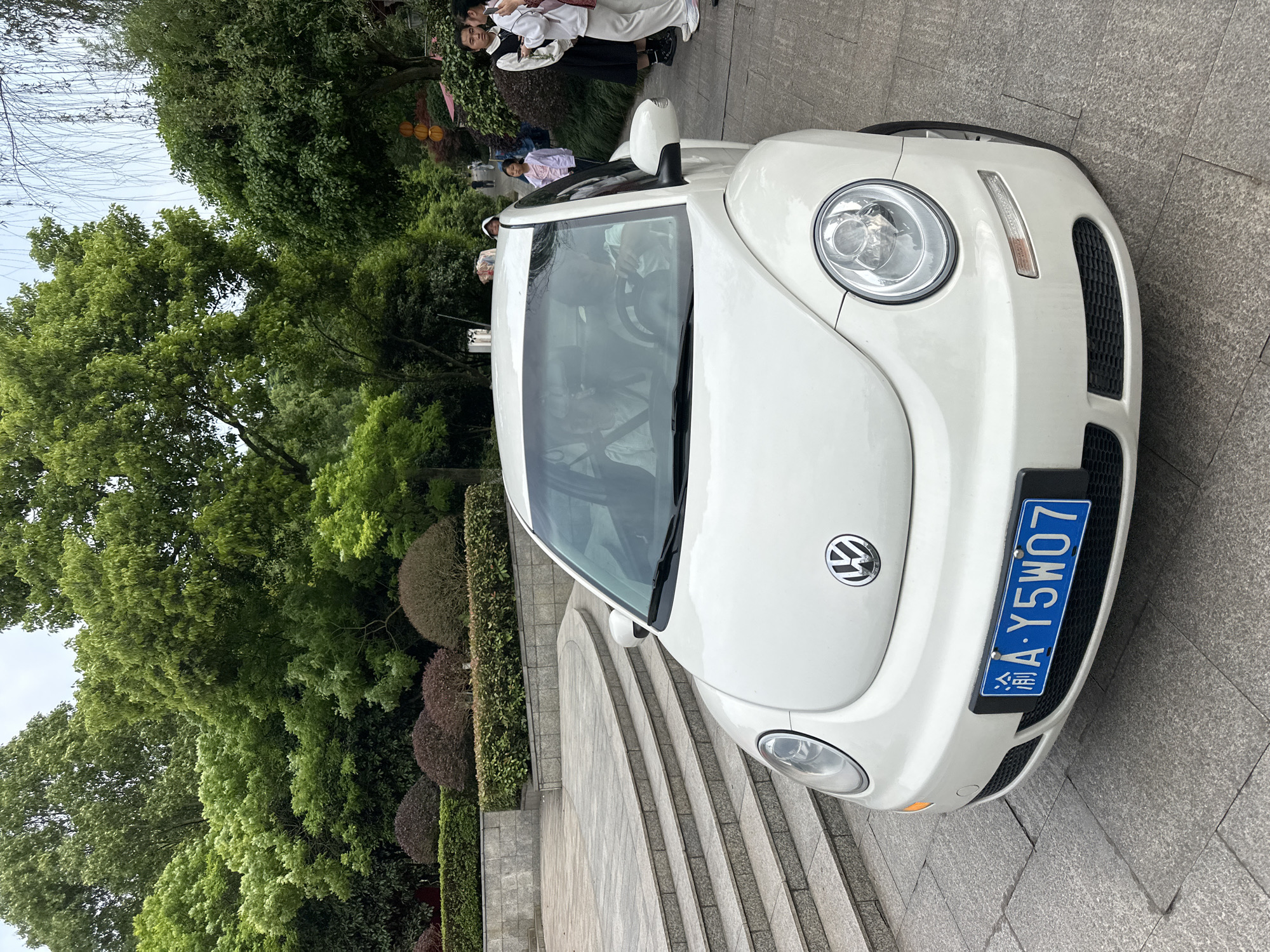 A white Volkswagen Beetle is parked on a stone staircase in an outdoor environment with lush greenery. The car has a blue license plate reading "渝A·Y5W07," indicating the car is registered in Chongqing. Several people are standing near the car, and they seem to be dressed in white uniforms. The background features trees and cultivated hedges.