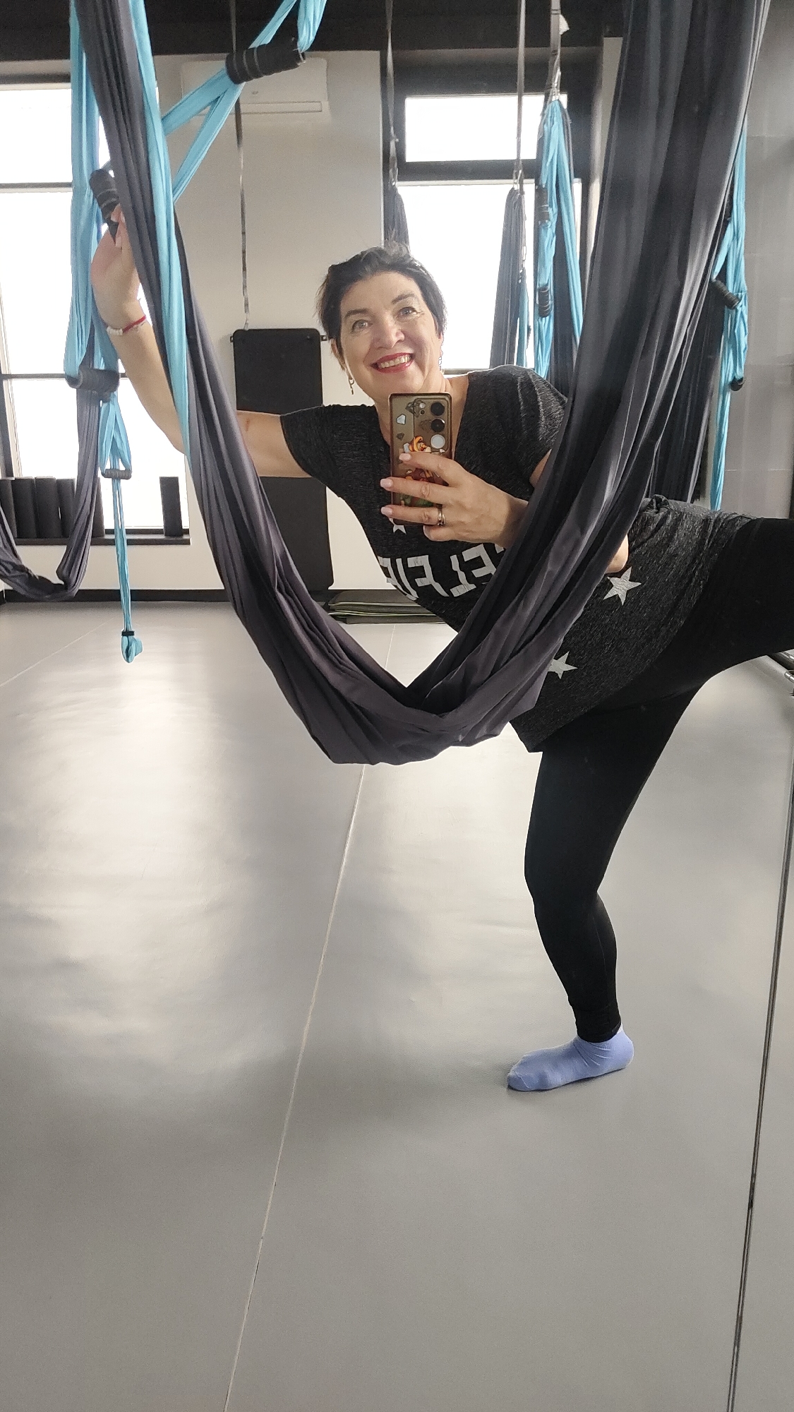 An adult woman with dark, short hair and a broad smile captures a mirror selfie in an aerial yoga studio in Krasnoyarsk, Russia. She is wearing a dark short-sleeved top adorned with white star patterns, black leggings, and light blue socks. Her right hand holds a smartphone in a patterned case, angled towards the mirrored wall. The woman is actively engaged with a dark grey aerial yoga hammock, with one leg extended to the side, showcasing a pose. Her shirt displays the partially visible white text "FEEL".
In the background, other light blue and dark grey hammocks hang from the ceiling, indicating the studio's purpose. The room features a light, reflective floor, dark walls, and large windows that allow bright daylight to illuminate the space. Stacked yoga mats are visible against a back wall, and an air conditioning unit is reflected high on the wall. The woman's happy expression suggests enjoyment of her activity.