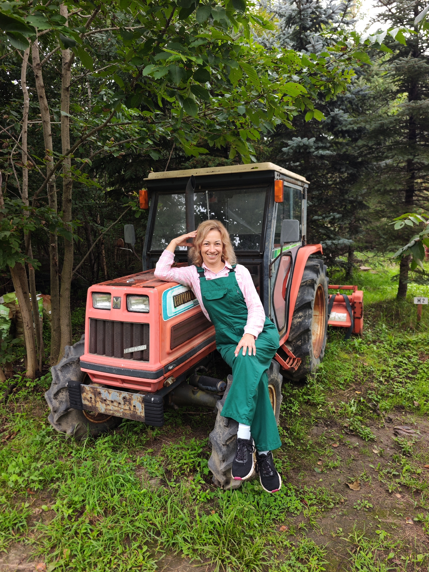 An outdoor, medium shot shows a smiling, middle-aged Caucasian woman with blonde, shoulder-length hair sitting on the front fender of an orange tractor. She is wearing a pink and white plaid long-sleeved shirt and green overalls. Her left arm is bent at the elbow with her hand resting on her head, and her right hand rests on her knee. Her legs are crossed at the ankles. The tractor has large rear tires and a prominent front grill. It is parked on a grassy area with trees and foliage in the background. There is a small red tiller attached to the back of the tractor. A sign with Cyrillic lettering and the number "2" is visible in the background to the right of the tractor. The scene appears to be in a rural or semi-rural setting, possibly within the vicinity of Solovej-Kljuch, Russia.