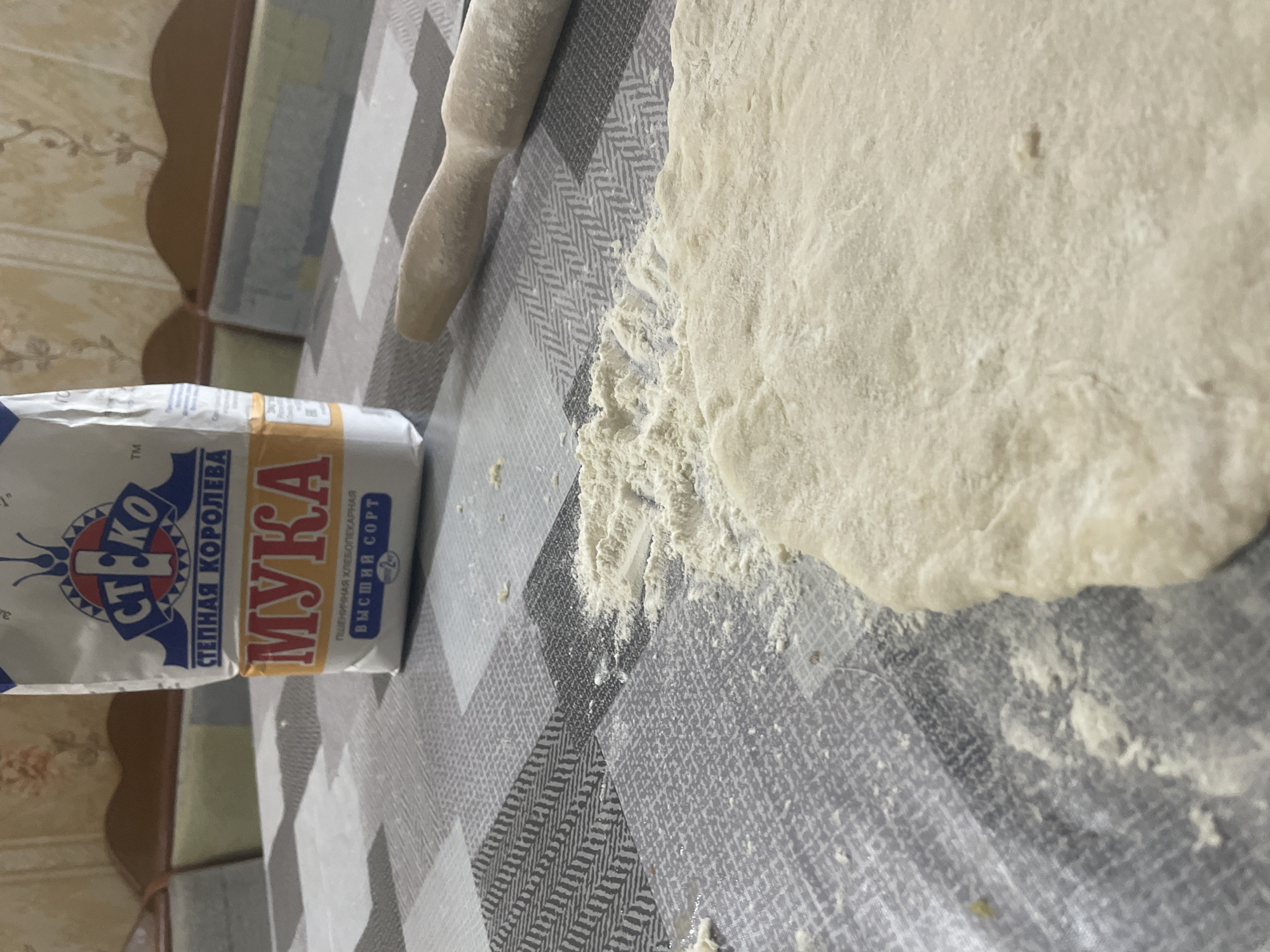 The image depicts a close-up, angled view of a table surface covered with a checkered tablecloth. On the left side, a white and blue bag of flour labeled "СТ. КО МУКА" is partially visible. The word "МУКА" is prominently displayed in large yellow lettering. Below this, text in smaller font reads "СТЕПНАЯ КОРОЛЕВА" and "ПШЕНИЧНАЯ ХЛЕБОПЕКАРНАЯ ВЫСШИЙ СОРТ".
To the right of the flour bag, a wooden rolling pin dusted with flour lies on the tablecloth. Adjacent to the rolling pin, a section of light-colored dough is spread out, also dusted with flour. Scattered flour is visible on the tablecloth around the dough and rolling pin, indicating recent activity.
The background shows a wall with a floral wallpaper pattern and a wooden shelf or furniture. The lighting suggests an indoor setting, possibly during the daytime. The overall scene implies food preparation, specifically baking, is in progress. The location context provided is Линёвка, Russia. There are no people visible in the image.
