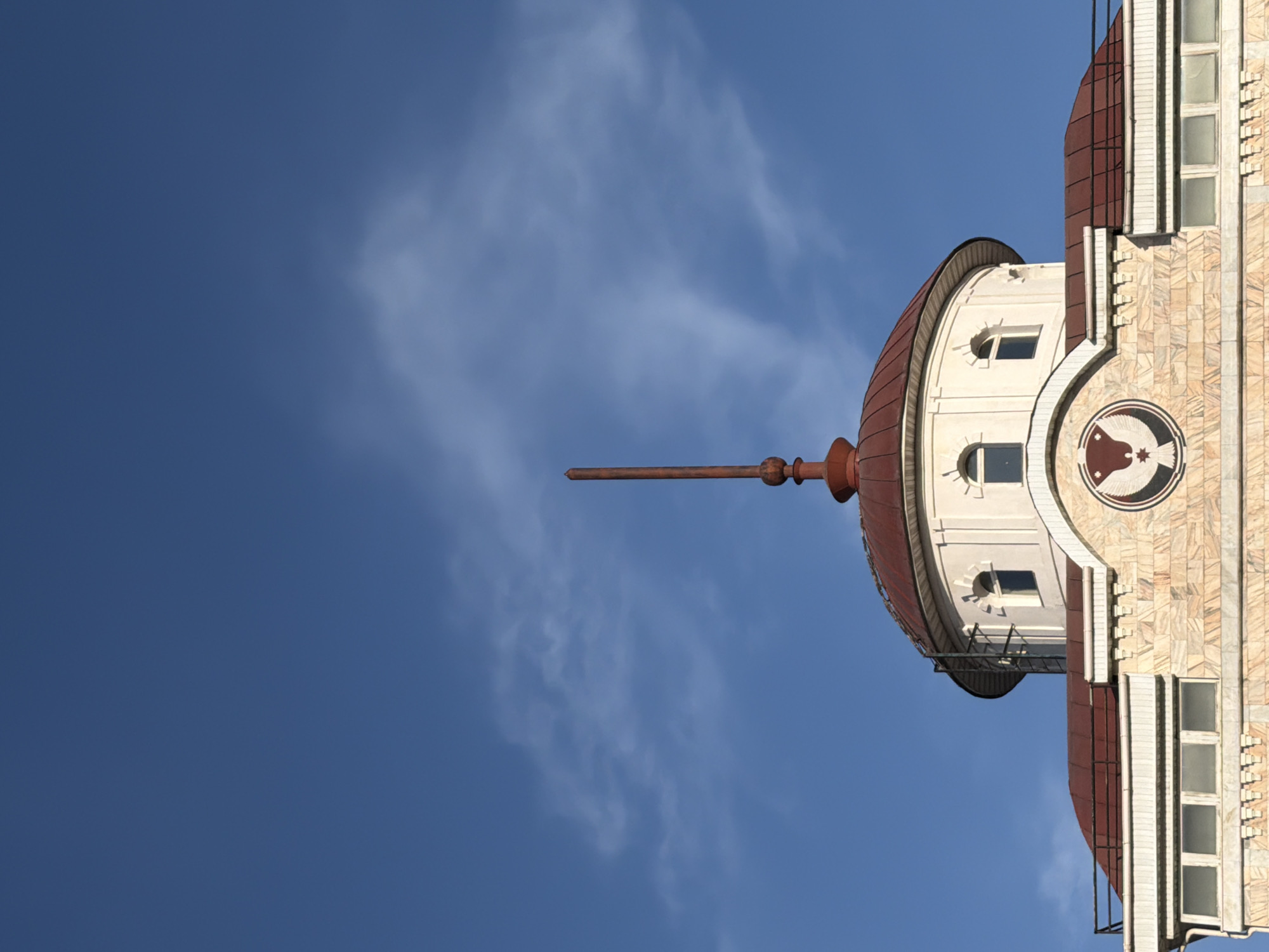 The image shows a building with a distinctive tower in Izhevsk, Russia. The tower features a reddish-brown dome with a prominent spire. The building's facade is a light beige color with a decorative, circular emblem containing a stylized image of a bird. The tower has small, arched windows. The background is a clear, blue sky with wisps of clouds. The image is oriented vertically.