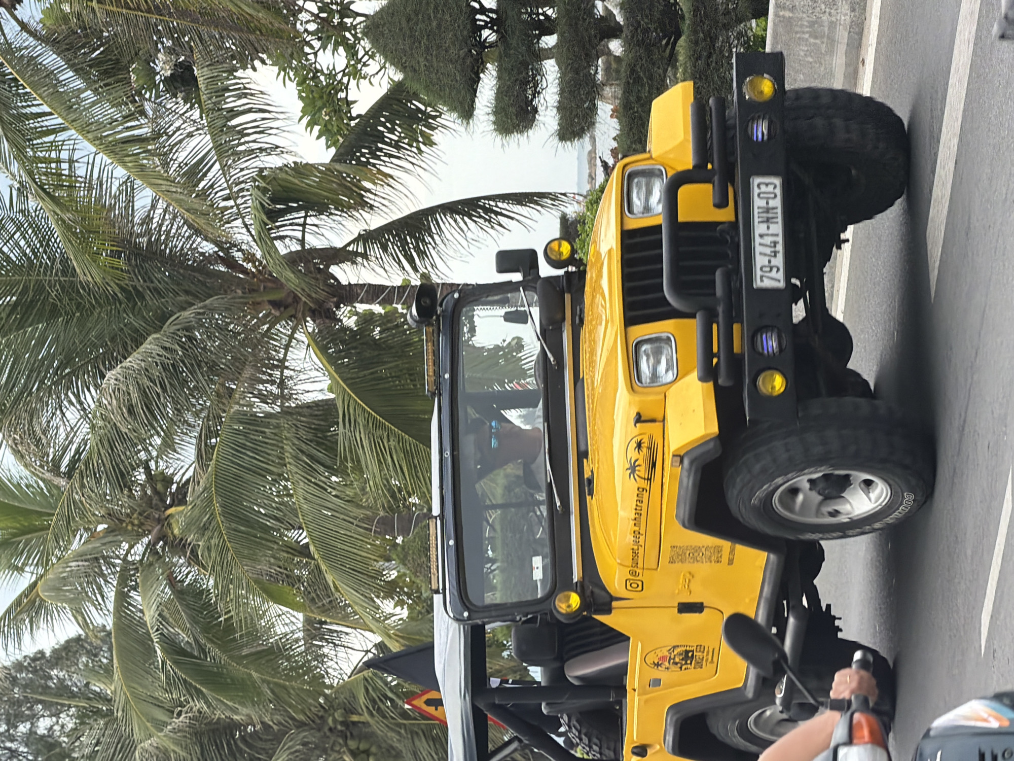 In Nha Trang, Vietnam, a bright yellow Jeep Wrangler, adorned with decals and rooftop lights, drives along a paved road. The Jeep features a black soft top and a black grille guard. A license plate, reading "79-441-NN-03", is mounted on the front. The driver, wearing sunglasses, is visible behind the windshield. Palm trees and lush green foliage line the road, with a glimpse of the ocean in the background. A black flag on a pole is mounted to the left side of the Jeep. A portion of a person's arm and a motorcycle are visible in the foreground on the left side of the image.