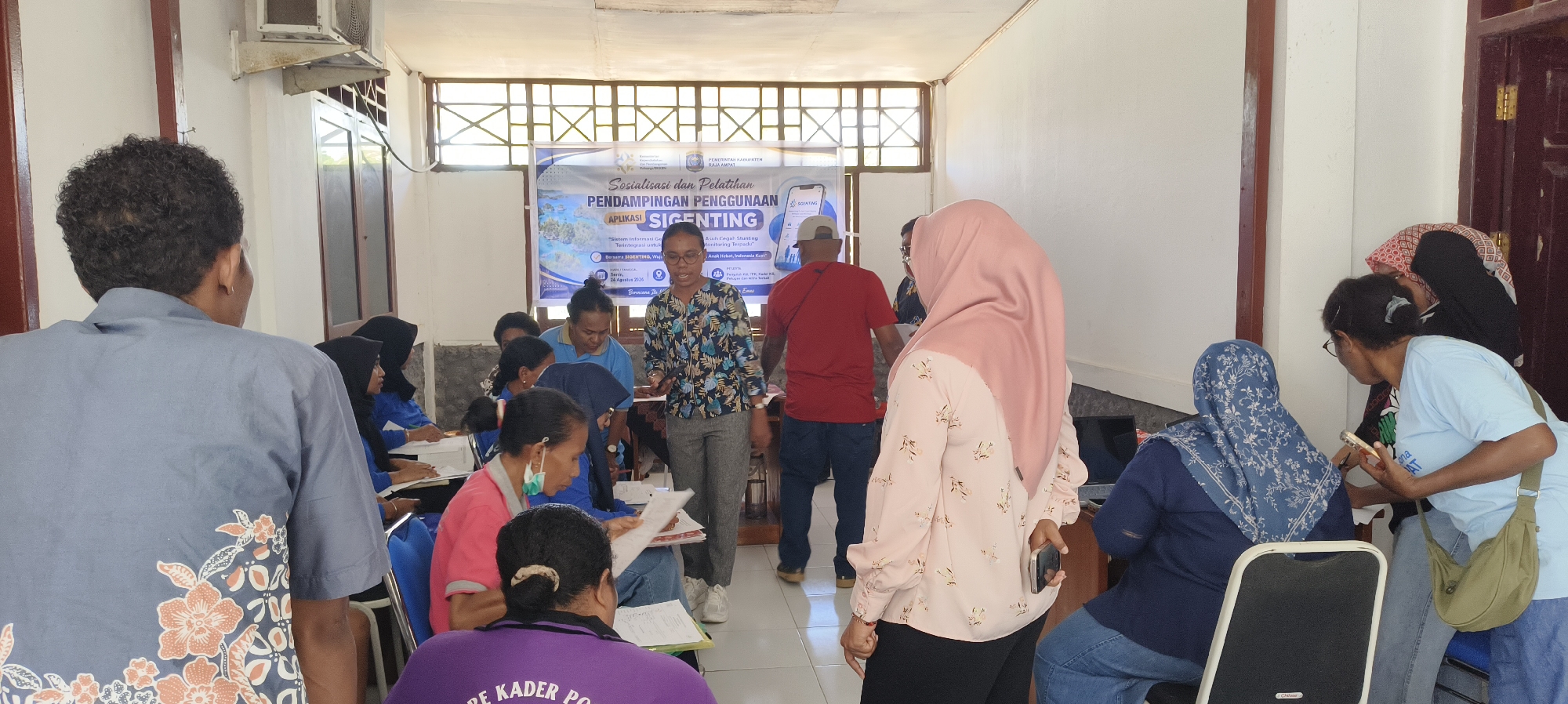 In an indoor community hall in Waisai, Indonesia, a group of about fifteen individuals, primarily women, are gathered for a meeting or training session. The room has white walls and a ceiling with exposed beams and fluorescent lighting. A large banner in the background, reading "SOSIALISASI DAN PELATIHAN PENDAMPINGAN PENGGUNAAN APLIKASI SIGENTING," indicates the event is a socialization and training for the "SIGENTING" application.
Several people are seated at tables, some with papers and pens, suggesting they are taking notes or participating in an activity. Others are standing and interacting. In the foreground, a person wearing a blue shirt with a floral pattern is visible from behind, facing away from the camera. To their right, a woman in a blue shirt and headscarf is seated at a table. A woman in a pink hijab and patterned shirt stands near the center, holding a smartphone. A man in a red t-shirt stands to her left, facing away from the camera, and another person in a blue shirt and headscarf is seated at a table. Further back, a woman in a floral patterned shirt and glasses is standing and speaking, gesturing with her hands, while others are seated around her. A person in a light blue t-shirt with a green messenger bag slung across their body is leaning over a table, looking at a phone. A few other individuals are partially visible, seated and standing, engaged in the session. The environment appears to be a functional, modestly furnished space.