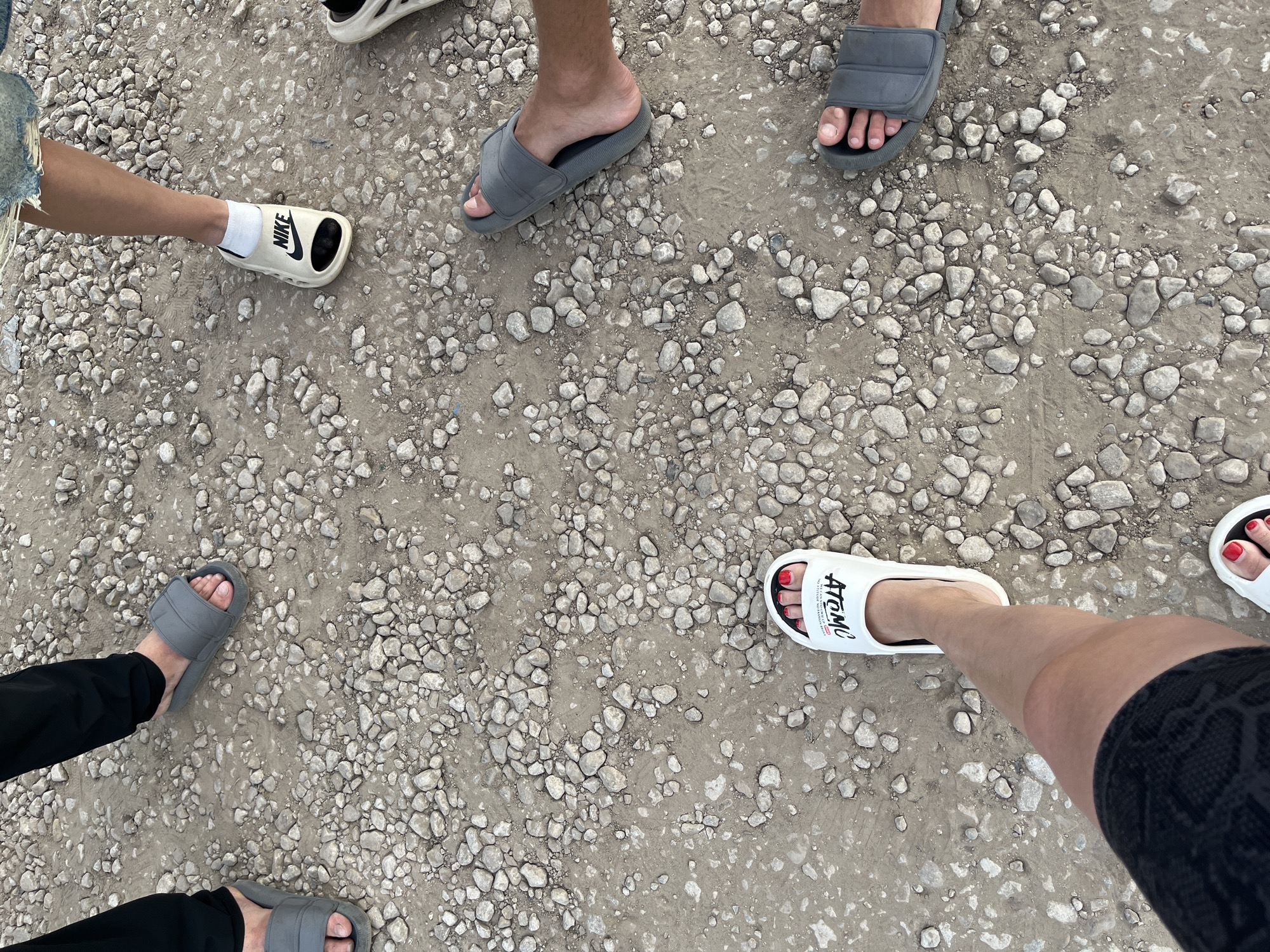 The image shows the feet of several individuals standing on a gravelly surface, likely a path or road in the city of Shumeyka, Russia. The ground is composed of small, light-colored stones and dirt.
One person is wearing black pants and gray slide sandals. Another individual is wearing white socks and cream-colored Nike slides. A third person is wearing denim shorts and gray slide sandals. A fourth person is wearing white slides with the word "ATOMO" printed in black, and another person is wearing similar white slides. The person wearing the "ATOMO" slides has bright red nail polish on their toes.
The scene appears to be a casual outdoor gathering. There is no indication of any specific event or organized activity. The lighting suggests it is daytime, possibly overcast. The focus is entirely on the lower extremities and footwear of the people present.