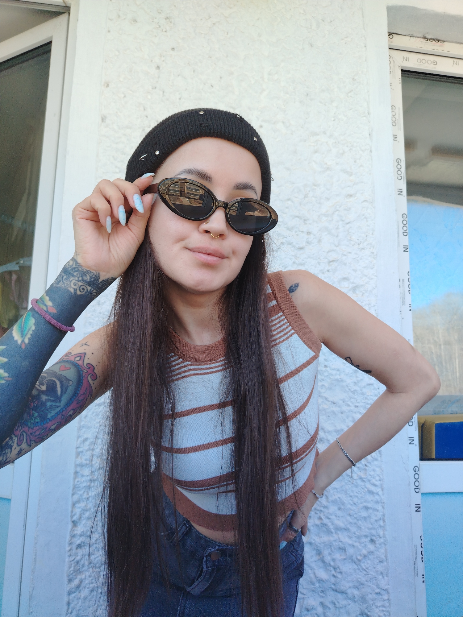 Here is a forensic description of the image:
**Scene:** The photograph appears to be taken outdoors, likely in a residential area, given the wall and window frame visible in the background. A woman is posing in front of a textured white wall with a window frame to the right.
**Subject:** The subject is a young woman with long, straight dark brown hair. She is wearing a black beanie, oval-shaped sunglasses, a horizontally striped brown and white tank top, and dark denim jeans. Her left arm is covered in a full-sleeve tattoo, and she has a nose ring. She is wearing a bracelet on her left wrist and a silver chain bracelet on her right wrist. Her fingernails are manicured with a light blue design.
**Action:** The woman is holding the frame of her sunglasses with her left hand, partially lowering them. She is looking directly at the camera with a slight smile. Her right hand is resting on her hip.
**Environment:** The background is a white, textured wall, and a window frame with "GOOD IN" printed several times along the vertical part of the frame. The window reflects the outside environment, which appears to be a sunny day.
There is no indication in the image the location is Noviy, Russia.