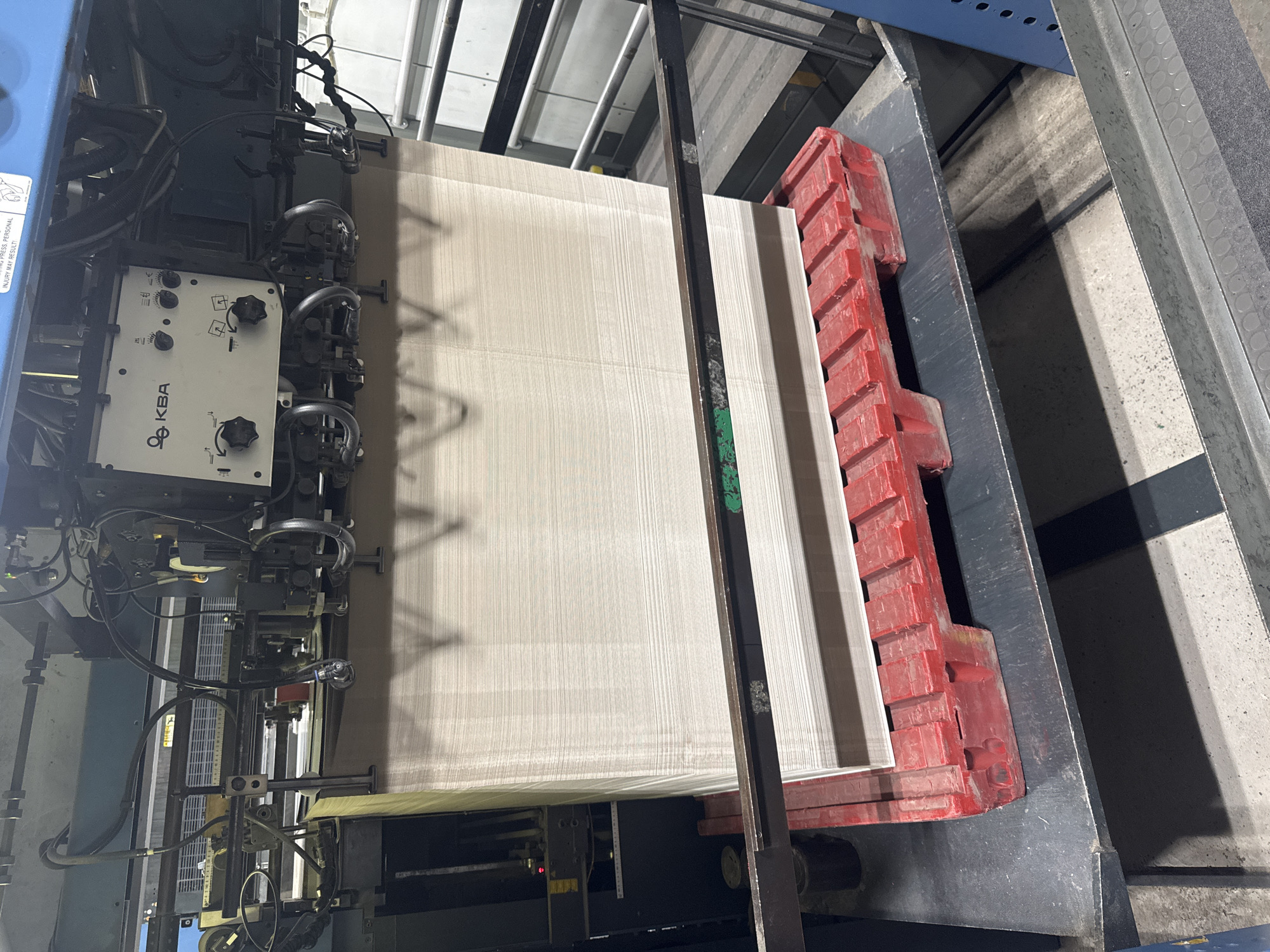 The image shows a large industrial printing press, likely in a factory setting in Jeffersontown, USA. The press is in operation, with a large stack of paper ready to be fed into the machine. The paper is light brown or tan and appears to be of a uniform thickness, stacked neatly on a red plastic pallet.
The press itself is complex, with various mechanical parts, wires, and control panels visible. One control panel, white with the logo "KBA," features several knobs and dials, suggesting manual adjustment capabilities. The overall scene is industrial and functional, with no people visible. The lighting appears to be artificial, typical of an indoor factory environment.
Notable details include the red plastic pallet holding the paper, the "KBA" branding on the control panel, and the neatly stacked paper. The environment appears clean but industrial, with concrete floors and metal structures visible in the background. The image captures a moment in the printing process, highlighting the machinery and materials involved in large-scale paper production or printing. There are no indicators of time of day or weather conditions.