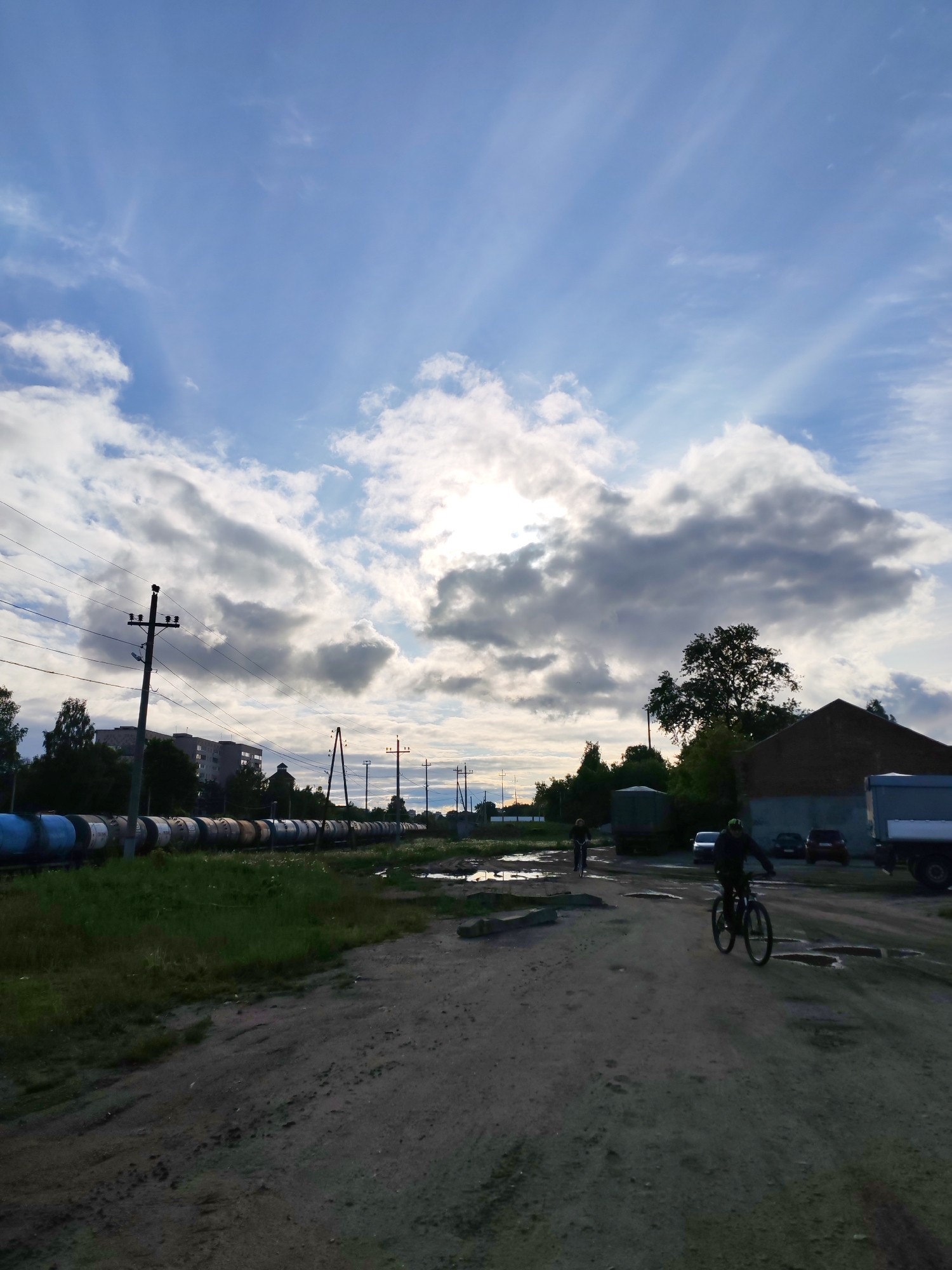 The scene depicts a partially overcast sky above an unpaved road in Bol'shie Mkhi, Russia. Two individuals are riding bicycles on the road, which is uneven and contains puddles. To the left, a line of tanker cars stretches along the edge of the road, bordered by greenery and power poles. In the background, buildings are visible among trees, along with parked vehicles near a building and trucks. The light is muted, and the overall environment suggests a working or industrial area.
