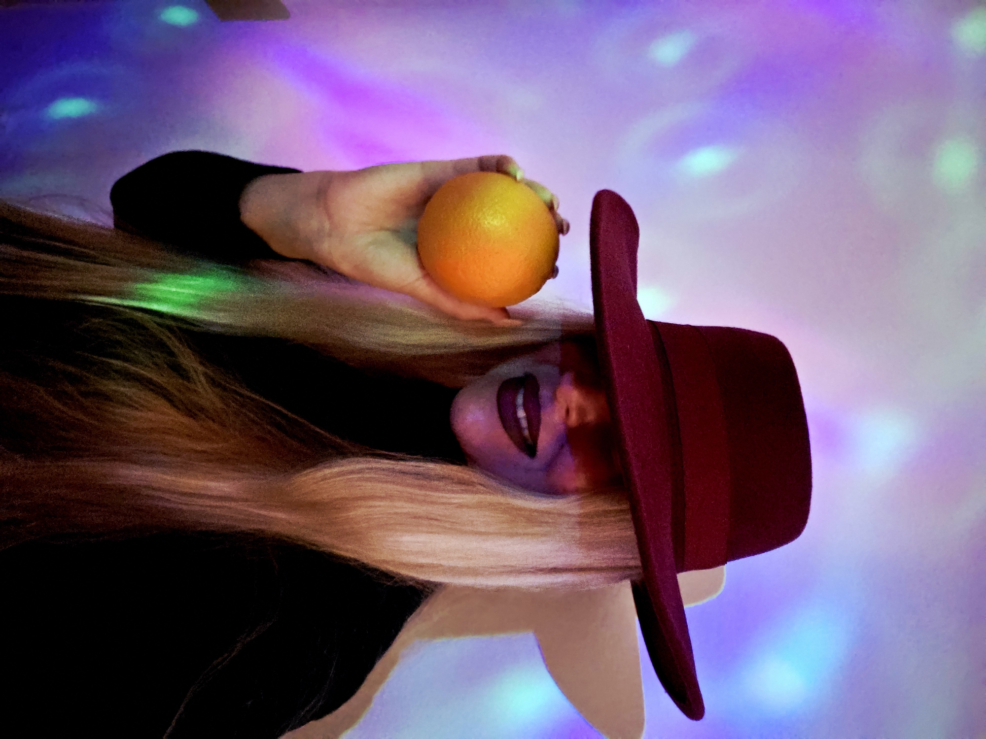 The image presents a stylized portrait of a person with long, flowing blonde hair. The individual is wearing a dark top and a wide-brimmed, dark red or burgundy hat. They are holding a bright orange in their right hand, positioned near their head.
The person's face is partially visible beneath the hat, featuring dark lipstick and a subtle smile or smirk. The lighting casts a purplish hue on parts of their skin, contributing to an artistic and somewhat surreal aesthetic.
The background is a vibrant, blurred expanse of colorful light, dominated by shades of purple, pink, and blue, with distinct, circular green and light blue reflections scattered across the surface. This luminous backdrop suggests an indoor setting, possibly a studio, a club, or a space with dynamic light projections, creating an atmosphere that is both playful and mysterious.
The image captures a posed moment, focusing on the interplay of color, light, and the subject's expression. No text is visible in the image. The setting implies a nighttime or artificially lit environment.