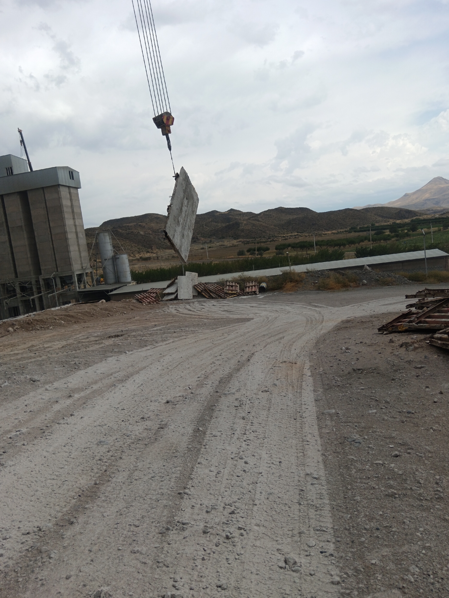 In Aragatsavan, Armenia, a construction site is depicted under a cloudy sky. A tall, gray industrial building stands to the left. A crane is actively lifting a large, flat concrete slab. Several large cylindrical silos are located near the building. In the foreground, a dirt road with tire tracks winds through the scene. In the background, rolling hills and distant mountains are visible. There are stacks of what appear to be construction materials on the right side of the image.