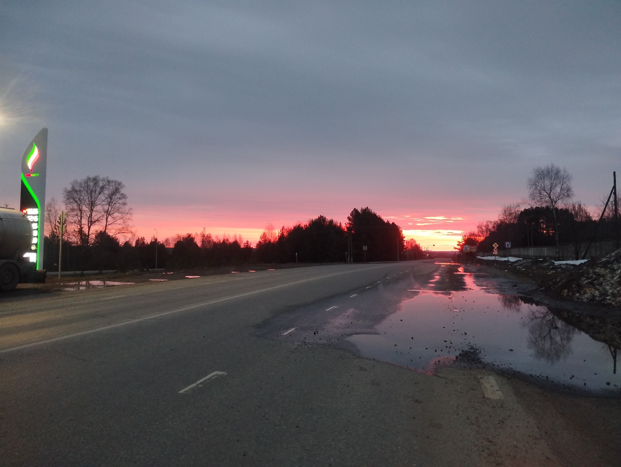 The image depicts a road leading into a sunset in Votkinsk, Russia. The road is asphalt, and there is a large puddle on the right shoulder, reflecting the colors of the sky. The sky is a mix of gray and pink, transitioning from dusk to night. On the left side of the road, there's a gas station sign with its logo illuminated. The roadside vegetation consists of trees, some still bare. Patches of snow are visible on the right. A truck is partially visible on the left side of the frame, near the gas station sign. A sign with a speed limit is positioned beyond the puddle.