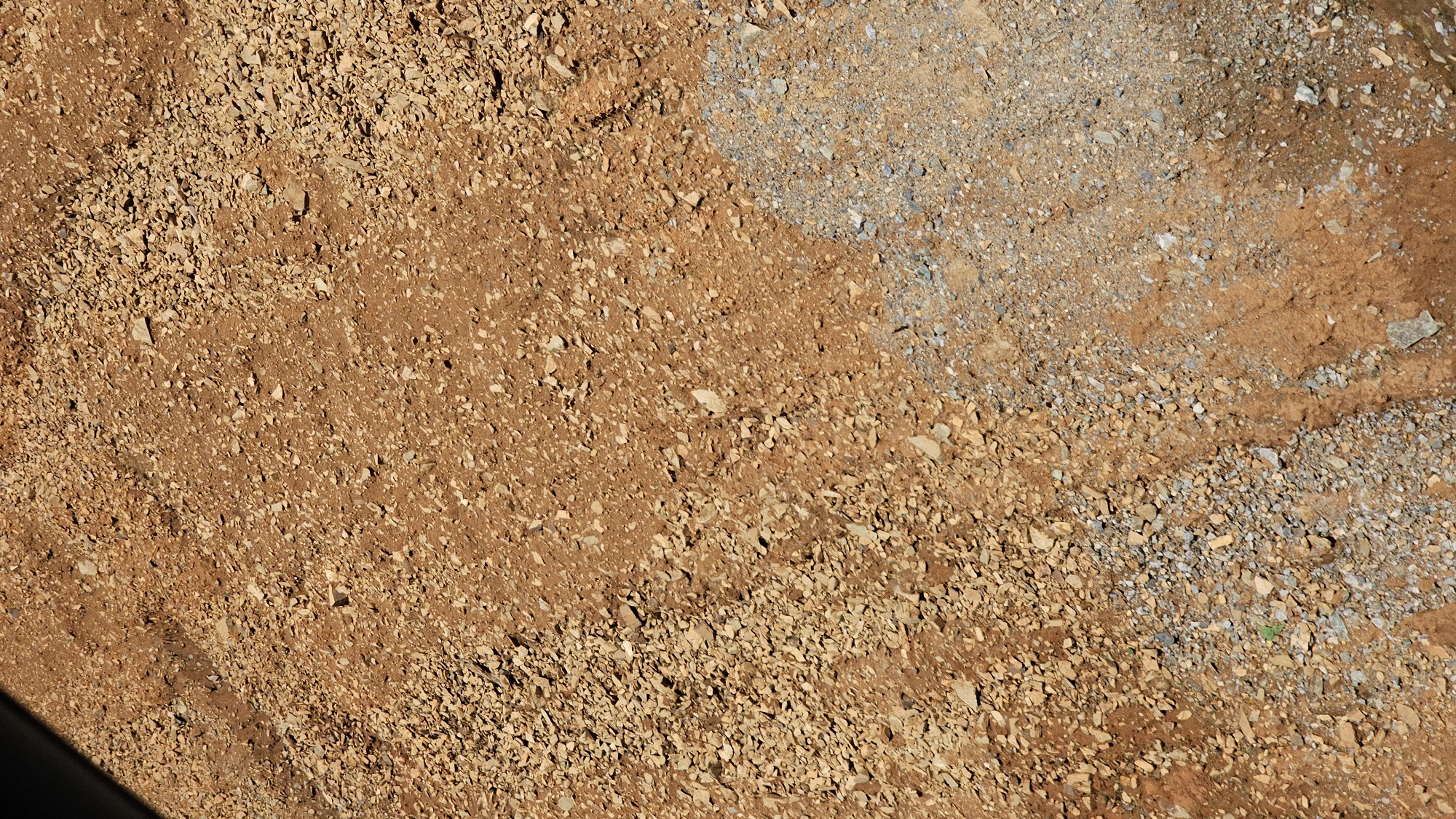 A close-up, top-down aerial view shows a texture of dry, reddish-brown earth mixed with gravel and small stones. A portion of the ground on the lower right is covered with a lighter-colored, grayish gravel. A dark shadow, likely from an overhead object or structure, is visible along the top left edge of the image. The scene is devoid of any people or identifiable landmarks.