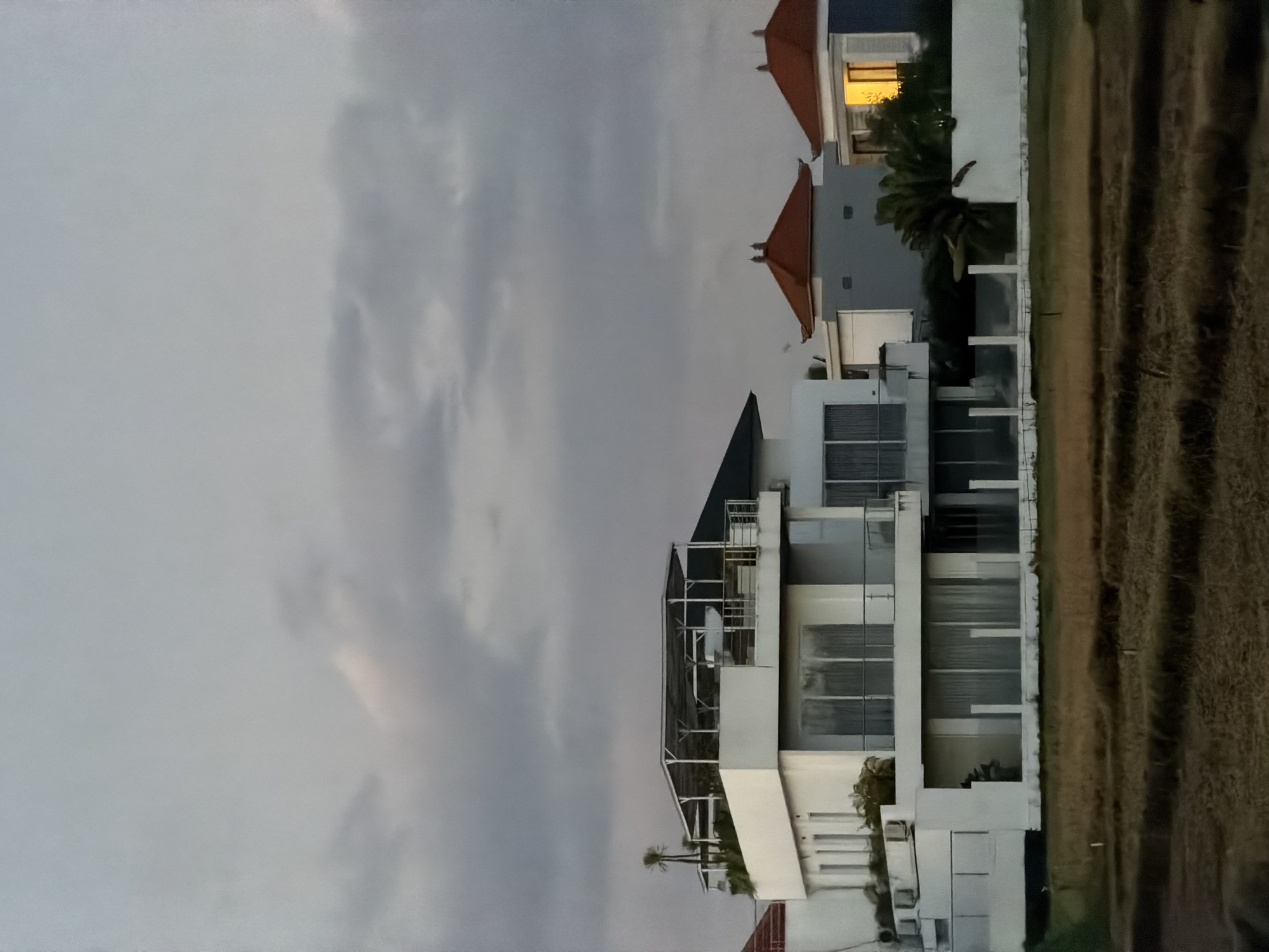 The image captures a scene in Mengwi, Indonesia, displaying a modern, multi-level white building with large windows and balconies. A dark, angled roof distinguishes one section. Adjacent to the building, a field of dry, golden vegetation stretches into the distance. The sky above is overcast with gray clouds, suggesting either dusk or a cloudy day. Other buildings with red-tiled roofs are also seen in the distance.
