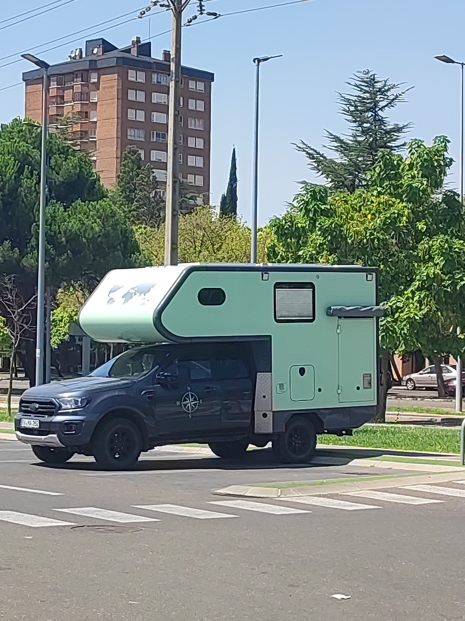 The image shows a light green camper van mounted on a dark gray pickup truck, parked on a city street in Palencia, Spain, near a pedestrian crossing. The camper van has a rounded front with a world map graphic, a side door, and a window. The pickup truck has black wheels and a compass decal on the door. In the background, there is a tall, brown brick building with many windows, lush green trees, a utility pole with power lines, and streetlights. The sky is clear and blue. The street appears to be a multi-lane road with grass and some vegetation bordering it.
