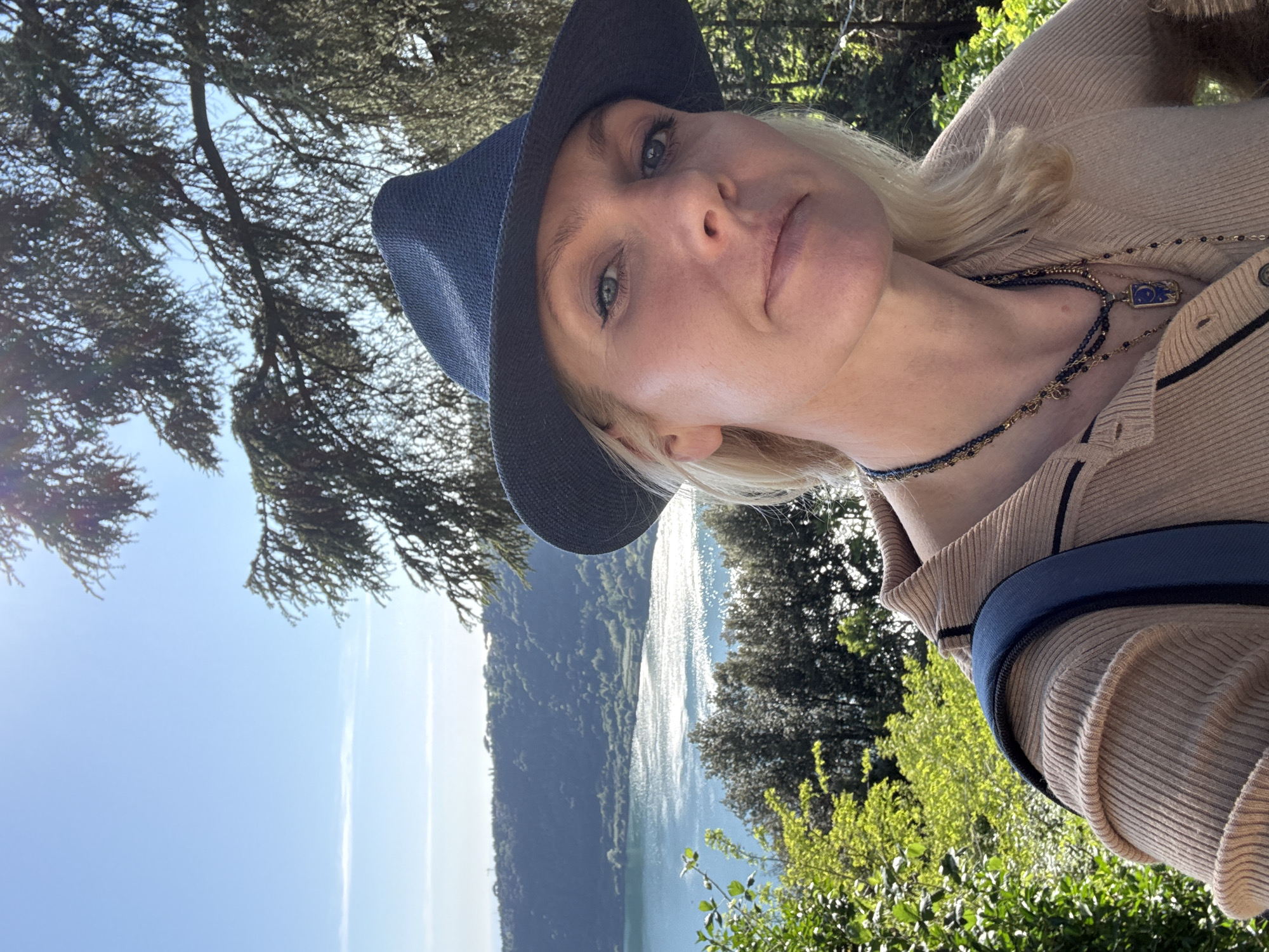 A woman with blond hair and a blue hat is facing the camera. She's wearing a tan sweater and a blue beaded necklace with a rectangular pendant. Behind her is a body of water reflecting sunlight, surrounded by green trees and a hill. The photograph was taken outdoors, likely in Nemi, Italy.