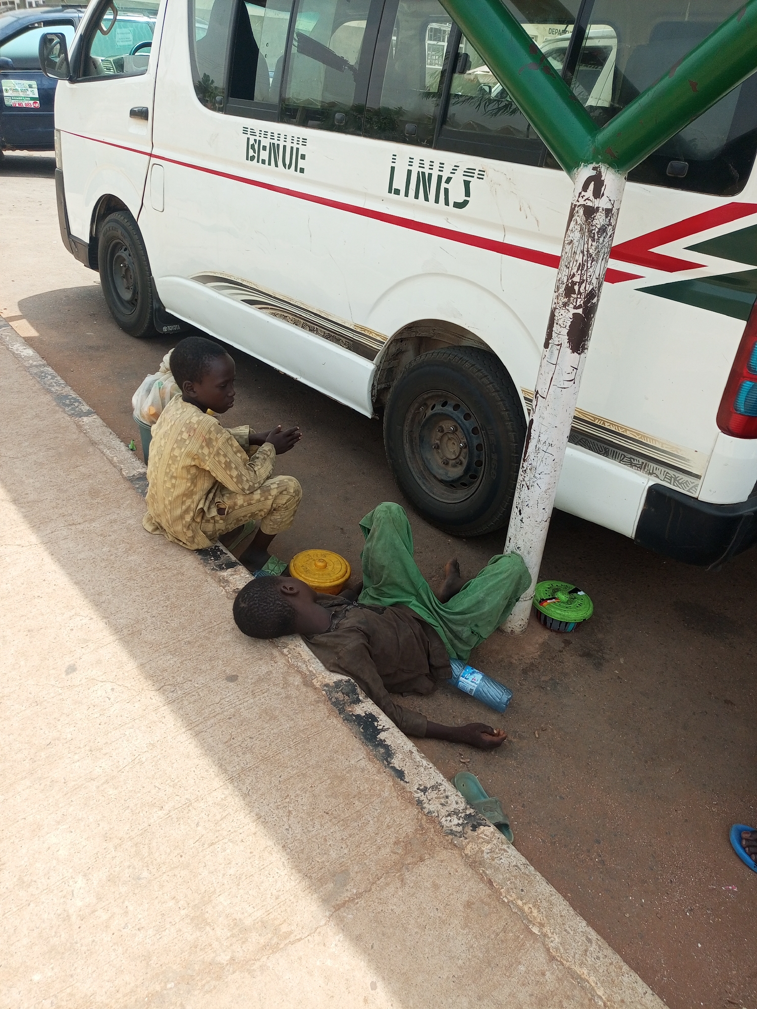 Two boys are located on a sidewalk next to a white "Benue Links" van. One boy is sitting cross-legged and dressed in patterned brown clothing. The other is lying on the ground wearing green pants and a dark shirt, with a plastic water bottle nearby. A yellow and a green object are visible near them. The scene appears to be in an urban area, possibly in the city of Gombe, Nigeria, based on the van's branding.