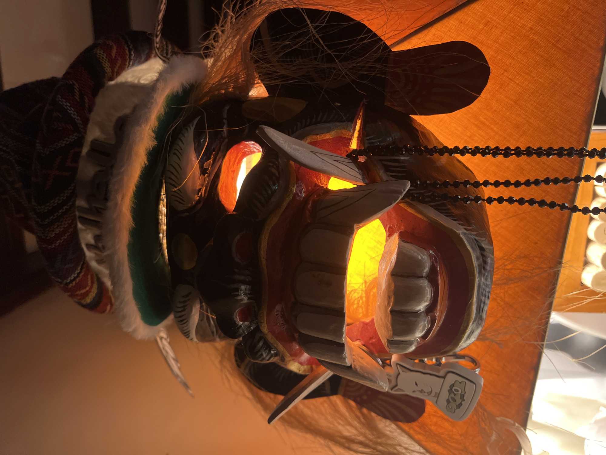 The image, rotated 90 degrees counter-clockwise, presents a close-up of an elaborately decorated mask, likely made of carved wood, illuminated from within. Bright orange and yellow light streams from the mask's eyes and wide-open mouth, highlighting its prominent white teeth and the intricate red and dark-colored facial features.
The mask is adorned with various decorative elements. A thick, white furry trim, backed by a band of green fabric, frames the upper left side of the mask as it appears in the frame. Above this trim, a patterned textile in shades of red, blue, and white with geometric designs is visible. Strands of light brown, fibrous material, resembling hair or straw, emerge from the mask's head area and sides. A black beaded chain hangs horizontally across the upper right portion of the mask.
Dangling from the lower right side of the mask's mouth is a small, white keychain shaped like a cartoon cat, with small green and yellow details beside it. The background consists of a warm, orange-brown textured surface, likely a lampshade, which glows softly, reinforcing the impression of an indoor setting during the evening or night. The mask appears to function as a unique light fixture or decorative object, situated in North Kuta, Indonesia. No legible text is visible other than possible indistinct markings on the keychain.
