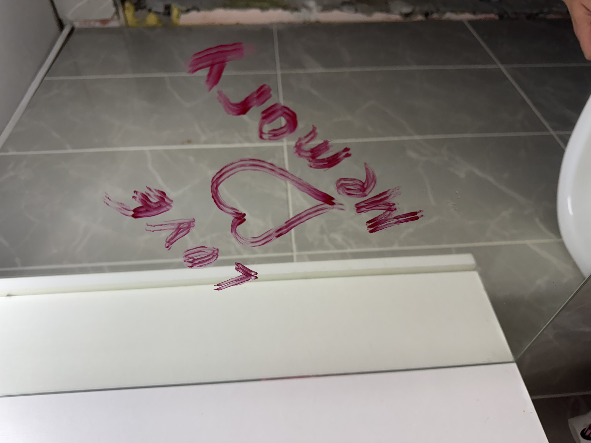 The image shows a bathroom mirror with lipstick writing. The words "I love you" are written on the mirror, accompanied by a heart symbol. The mirror is situated above a white vanity. The background reveals grey tiles and unfinished wall patching, suggesting a bathroom undergoing renovation. The location is unspecified, and there are no identifiable objects or people to tie it to Chaikovsky, Russia.