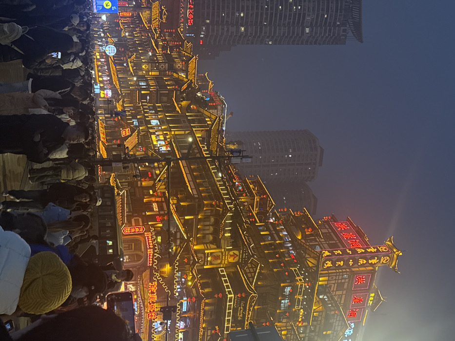 The image depicts a densely crowded nighttime urban scene in Yuzhong District, Chongqing, China, specifically focused on the illuminated Hongyadong complex.
**Environment:** The setting is an urban street or plaza at night, under a dark, hazy sky. The ground surface appears paved. Artificial light sources dominate the scene, including numerous golden architectural lights, bright red neon signs, and streetlights.
**Structures:** The most prominent subjects are the traditional multi-tiered stilt-house (diaojiaolou) style buildings of Hongyadong, occupying the right and central portions of the upper frame. These structures are extensively outlined and lit with warm golden lights, featuring numerous red Chinese lanterns hanging from eaves. Multiple levels display bright red neon signs with Chinese characters, notably "洪崖洞" (Hongyadong) and "重庆小天鹅" (Chongqing Little Swan). In the background, to the left and center, several modern, grey high-rise buildings with illuminated windows contrast with the traditional architecture. A white traffic sign with blue directional arrows is visible on the far left. A vehicle, possibly a bus, is partially visible mid-frame on the right.
**People:** A dense crowd of approximately one hundred individuals occupies the lower half to two-thirds of the image. The crowd consists primarily of adults of various ages, casually dressed in jackets, sweaters, and some with hats, indicating cooler weather. Individuals are engaged in various actions: standing, walking, observing the illuminated buildings, and frequently holding up smartphones to capture photographs or videos of the scene. Interactions are consistent with a large gathering of tourists or leisure-seekers. No specific organized activity or conflict is discernible within the crowd.