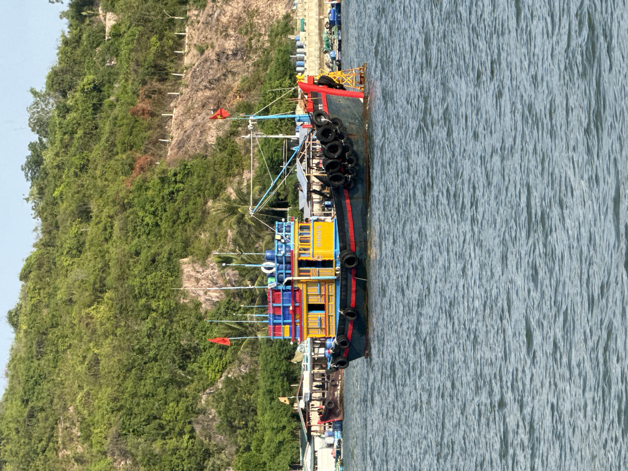 A colorful fishing boat with red, yellow, blue, and black markings is docked in the water. The boat has multiple flags and is equipped with tires along its hull for protection. In the background, there is a lush green hillside covered in trees. Several other boats and structures are visible along the shoreline. A person can be seen standing on the deck of the fishing boat, and another person is visible on a smaller boat in the distance. The water appears calm with small ripples. The scene suggests a harbor or coastal area in Nha Trang Ward, Vietnam.