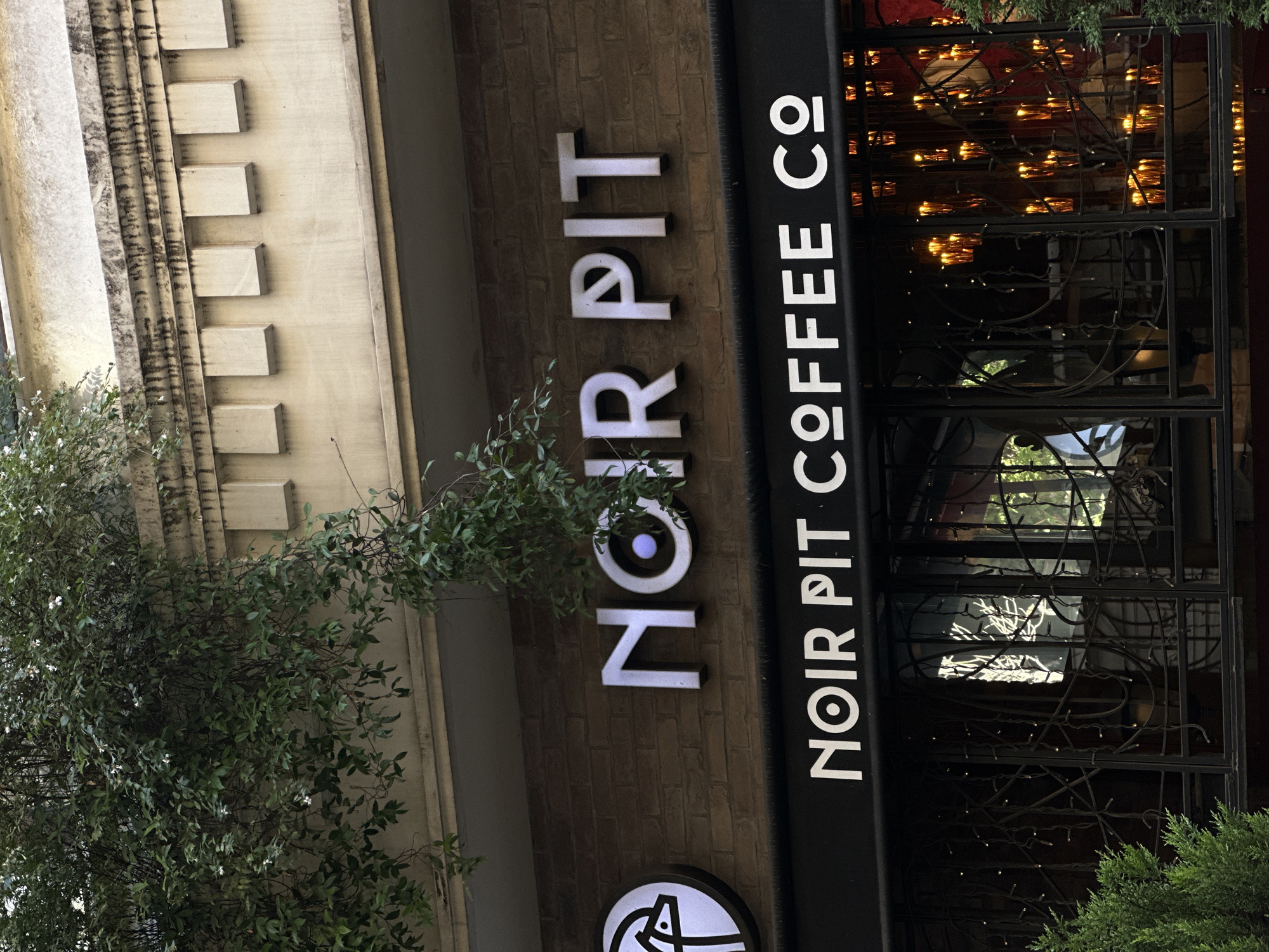 The image shows the exterior of a coffee shop named "NOIR PIT COFFEE CO" located in Beyoğlu, Turkey. The signage is prominently displayed above the entrance. The upper signage reads "NOIR PIT" in large, illuminated white letters against a brick wall. Below this, a black awning displays "NOIR PIT COFFEE CO" in smaller white lettering. The entrance features decorative black metalwork and is illuminated by numerous small, amber-colored lights, suggesting an evening or interior lighting display. A green bush or tree is visible in the lower left corner, and foliage also drapes from above the building's facade. The architectural style suggests a somewhat older building, with decorative elements on the upper structure. The overall scene depicts a commercial establishment in an urban setting.