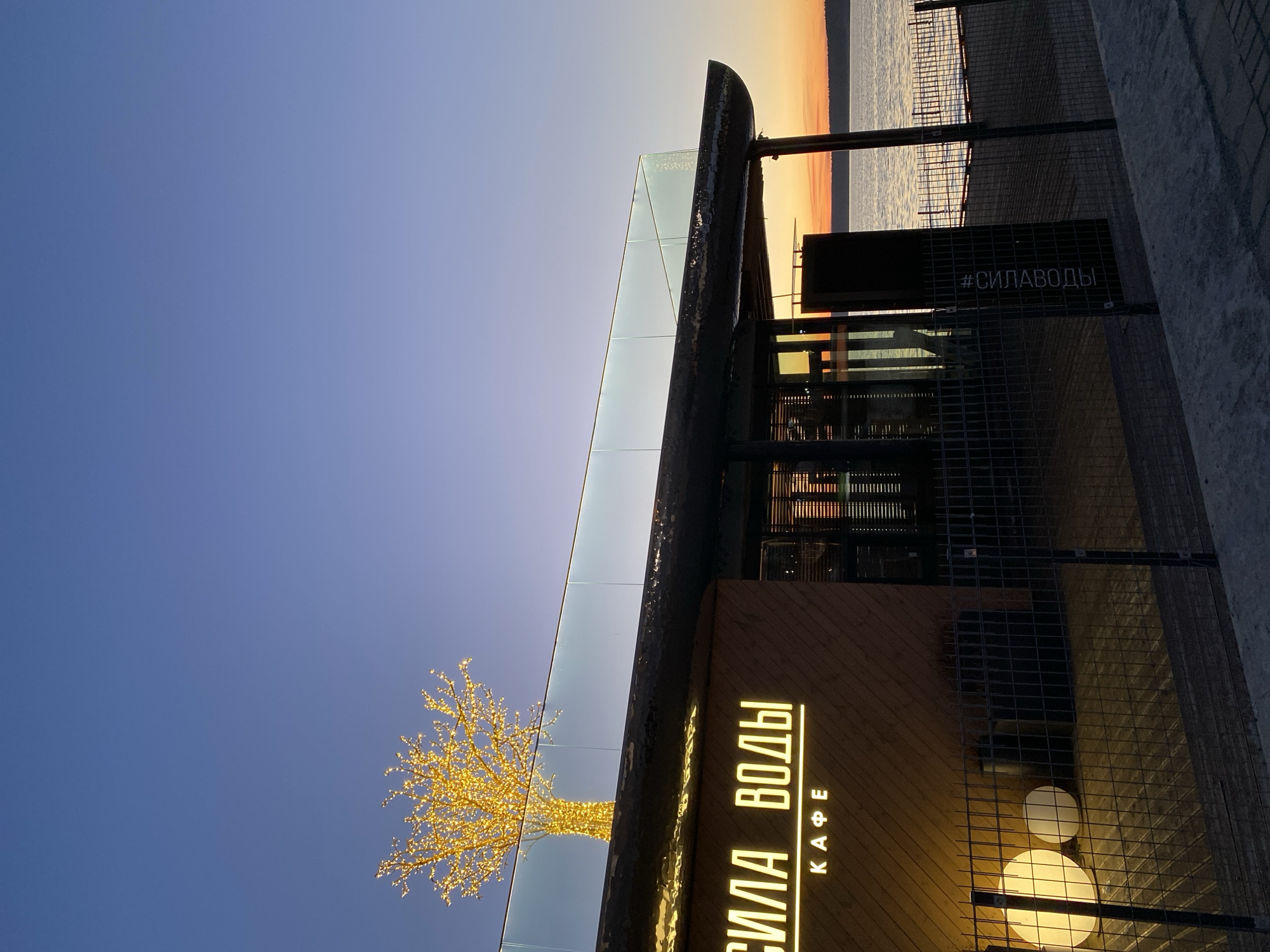 Here's a concise description of the image:
The photo shows a building with the illuminated words "СИЛА ВОДЫ КАФЕ" (Sila Vody Cafe) and a glowing tree-like structure next to it. A sign with "#СИЛАВОДЫ" is also visible. The sky is a gradient of blue to orange at the horizon. The location is likely Votkinsk, Russia, based on the cafe's name ("Sila Vody" meaning "Power of Water"), which aligns with Votkinsk's reservoir and water-related context. The photo appears to be taken at dusk or twilight, given the light levels and sky color.